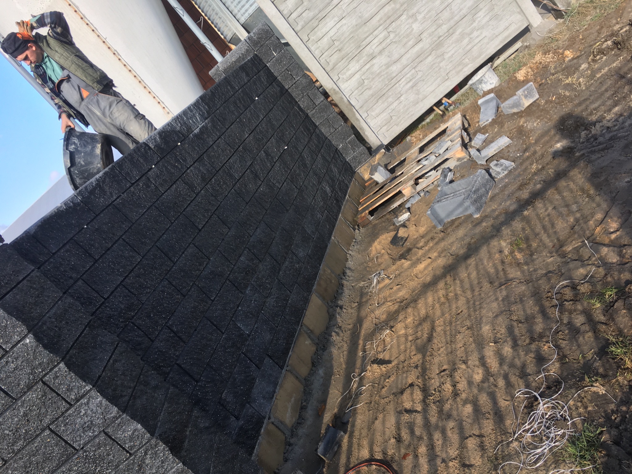 Budowa muru z czarnych, prostokątnych bloczków betonowych, widoczny pracownik z wiadrem, paleta z elementami, ziemia i fragment ogrodzenia w tle.