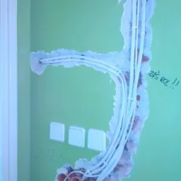 Ściana w trakcie kładzenia instalacji elektrycznej, widoczne przewody w białej osłonie wychodzące z puszki, trzy włączniki światła zamontowane obok siebie, całość na tle zielonej ściany z napisem...