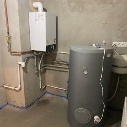 HYDROSTREFA ANDRZEJ SZCZĘSNY - Nowy, biały piec gazowy z podłączonymi miedzianymi rurami i szary zbiornik na wodę w surowym pomieszczeniu z betonowymi ścianami i podłogą zabezpieczoną niebieską taśmą.