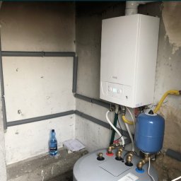 HYDROSTREFA ANDRZEJ SZCZĘSNY - Nowy, biały piec gazowy wiszący na ścianie, z widocznymi podłączeniami, rurami w szarej izolacji, niebieskim zbiornikiem wyrównawczym i butelką wody mineralnej w tle.