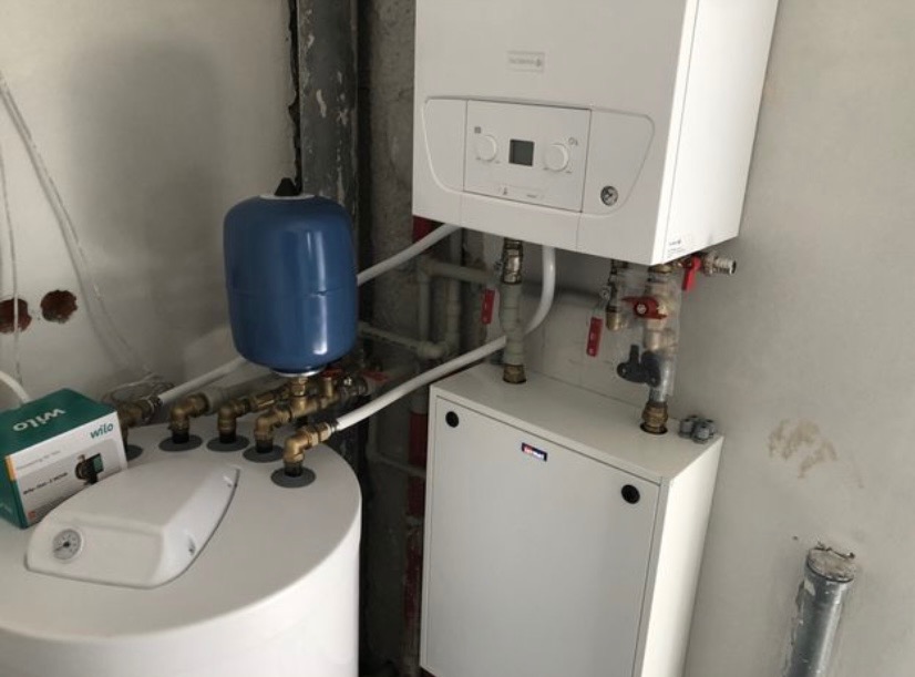 Nowoczesny, biały piec gazowy kondensacyjny zamontowany obok zbiornika na wodę z niebieskim naczyniem wzbiorczym, widoczne rury i zawory, surowy stan wykończenia ściany.