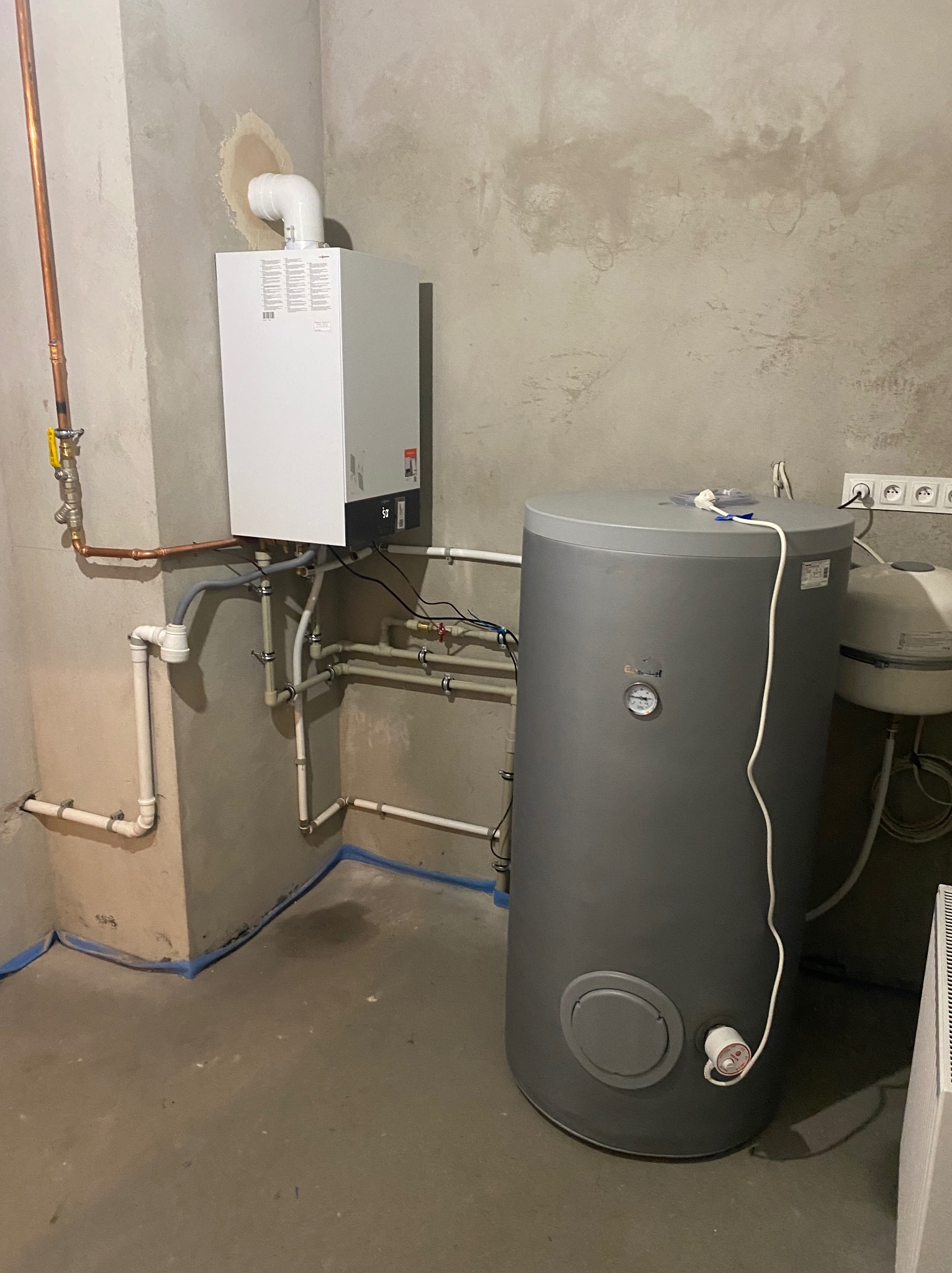 Nowy, biały piec gazowy z podłączonymi miedzianymi rurami i szary zbiornik na wodę w surowym pomieszczeniu z betonowymi ścianami i podłogą zabezpieczoną niebieską taśmą.