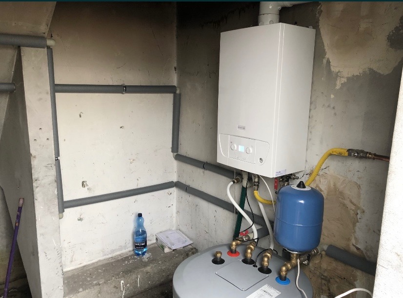 Nowy, biały piec gazowy wiszący na ścianie, z widocznymi podłączeniami, rurami w szarej izolacji, niebieskim zbiornikiem wyrównawczym i butelką wody mineralnej w tle.