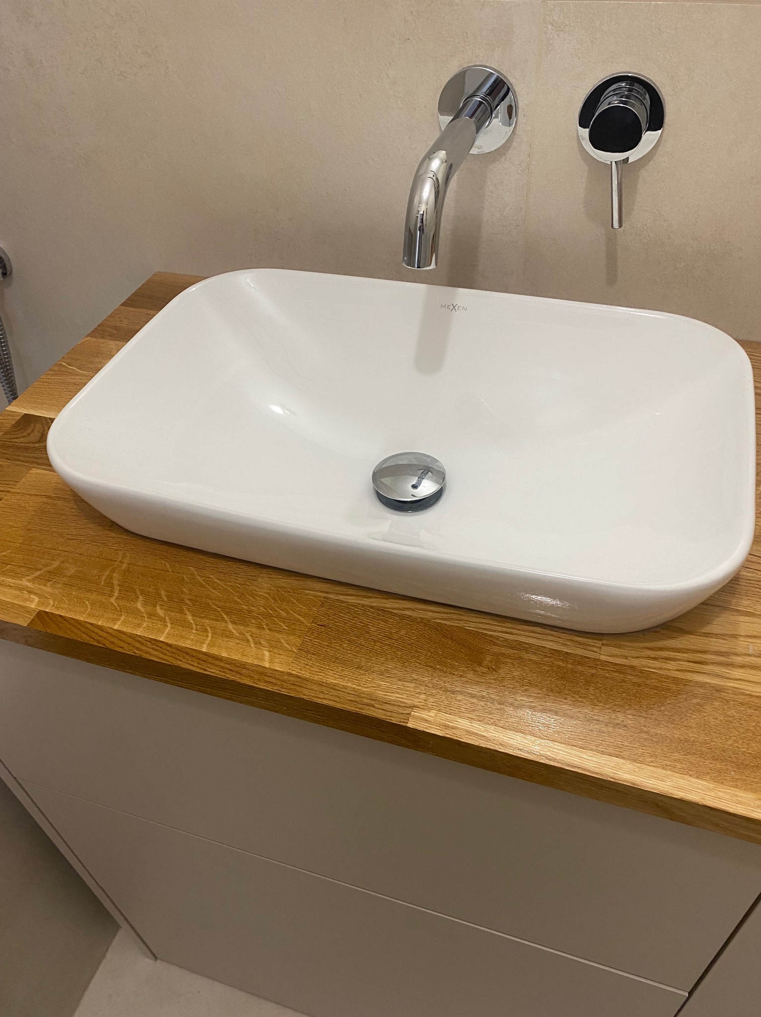 Biała, prostokątna umywalka nablatowa marki Mexen z chromowaną baterią ścienną i korkiem automatycznym, zamontowana na blacie z drewna dębowego, z szafką w kolorze taupe.
