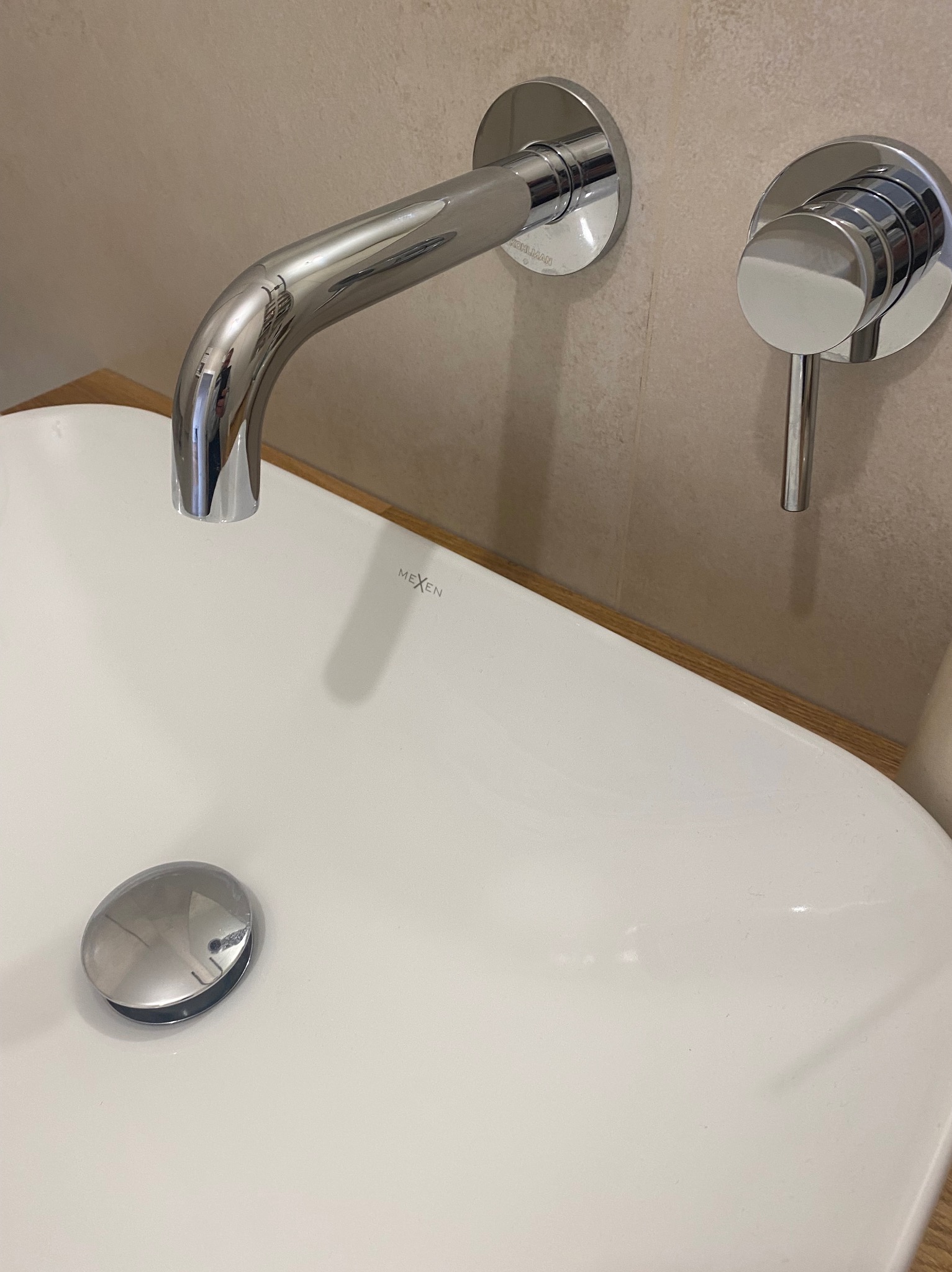 Bateria umywalkowa ścienna marki Kohlman i Mexen zamontowana na umywalce nablatowej, z widocznym korkiem automatycznym i beżową płytką w tle.