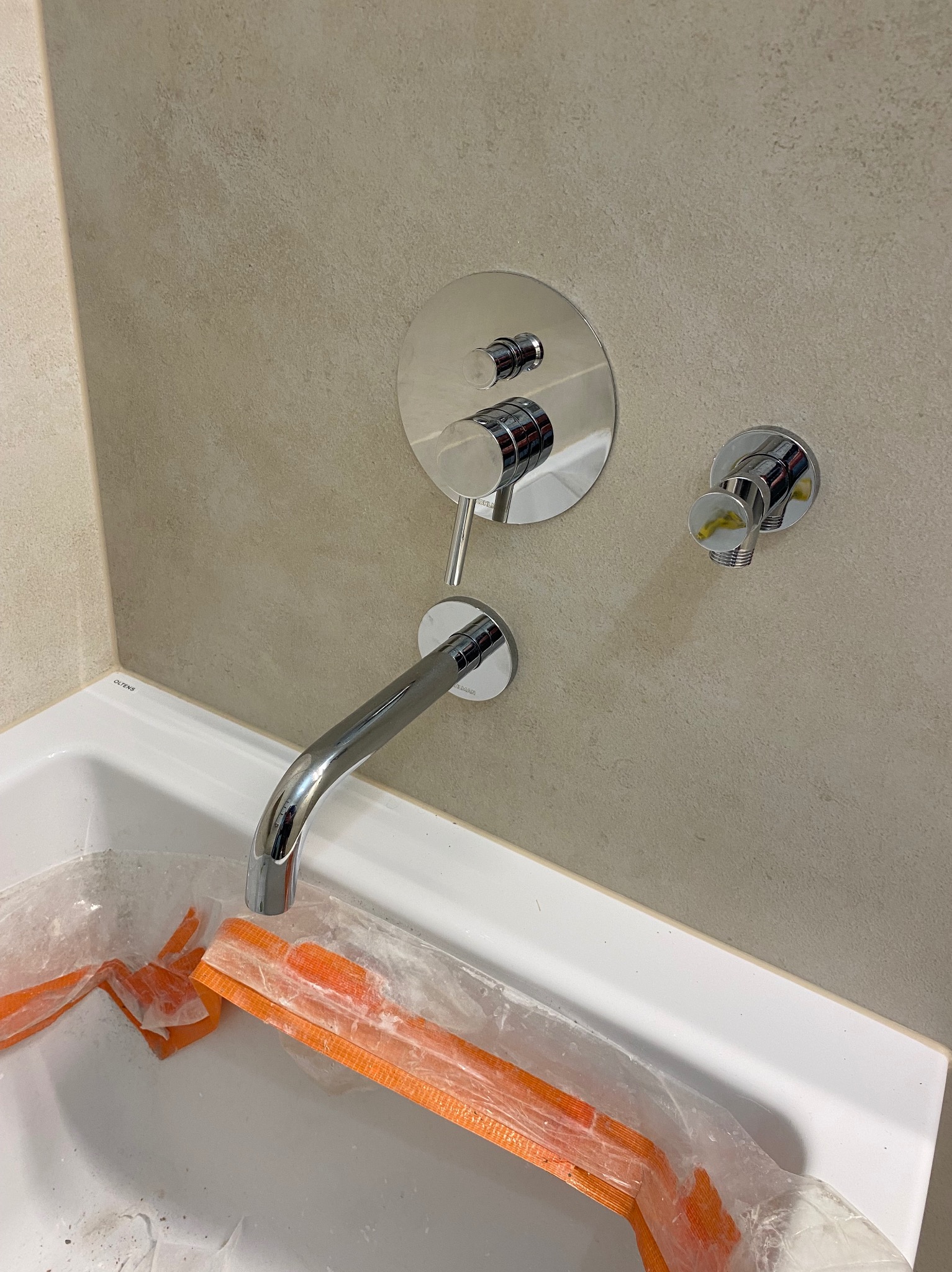 Bateria umywalkowa chromowana, montowana na ścianie, z okrągłą rozetą i wylewką, widoczna nad białą umywalką zabezpieczoną folią i pomarańczową taśmą uszczelniającą.