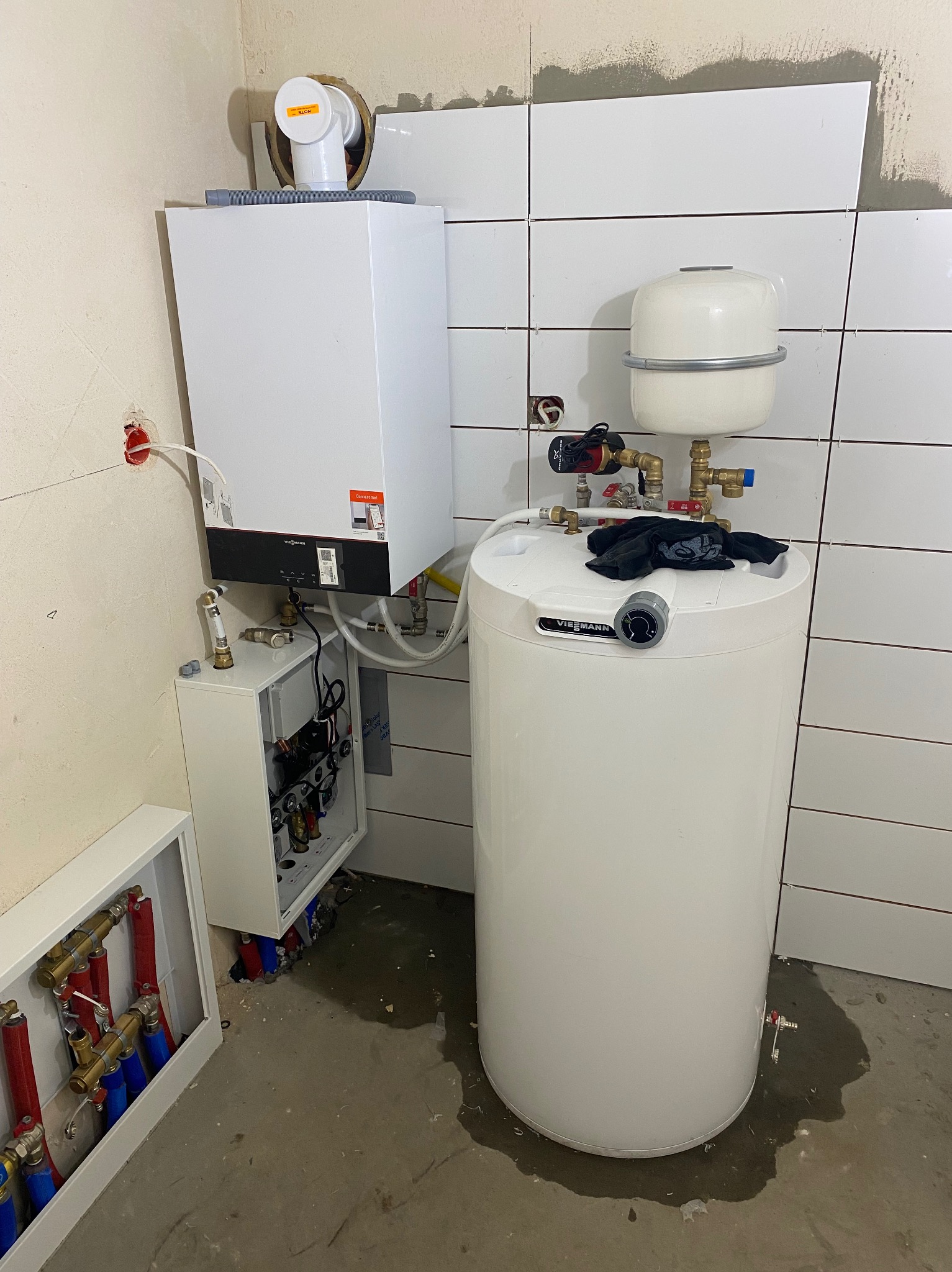 Nowoczesny, biały piec gazowy Viessmann z otwartym panelem sterowania, zbiornikiem na wodę i rozdzielaczem podłogówki podczas montażu w surowym wnętrzu z częściowo położonymi białymi kaflami.