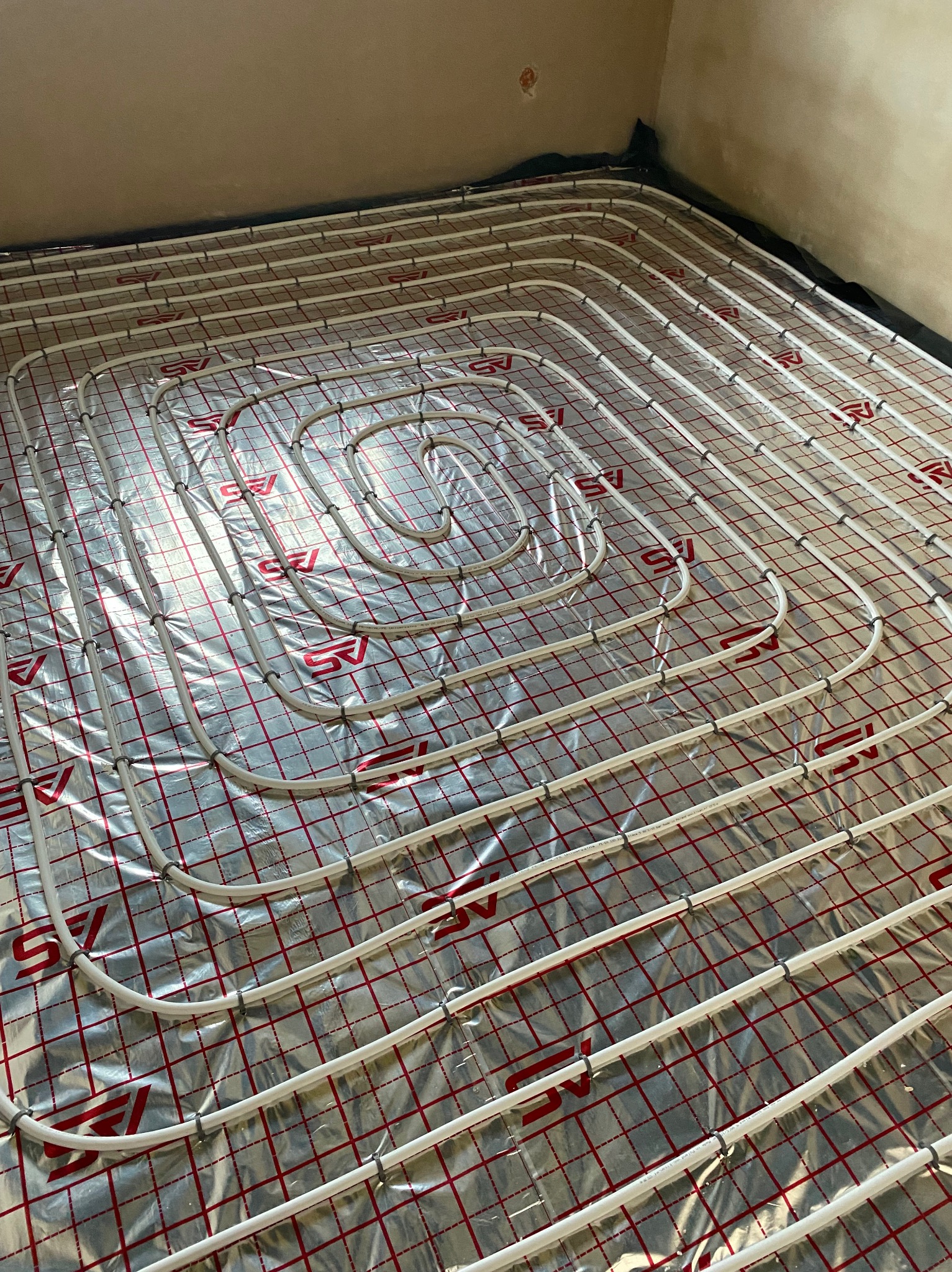 Białe rury ogrzewania podłogowego ułożone spiralnie na folii aluminiowej z czerwoną siatką, zamocowane plastikowymi klipsami, widok z góry, przygotowane do zalania wylewką.