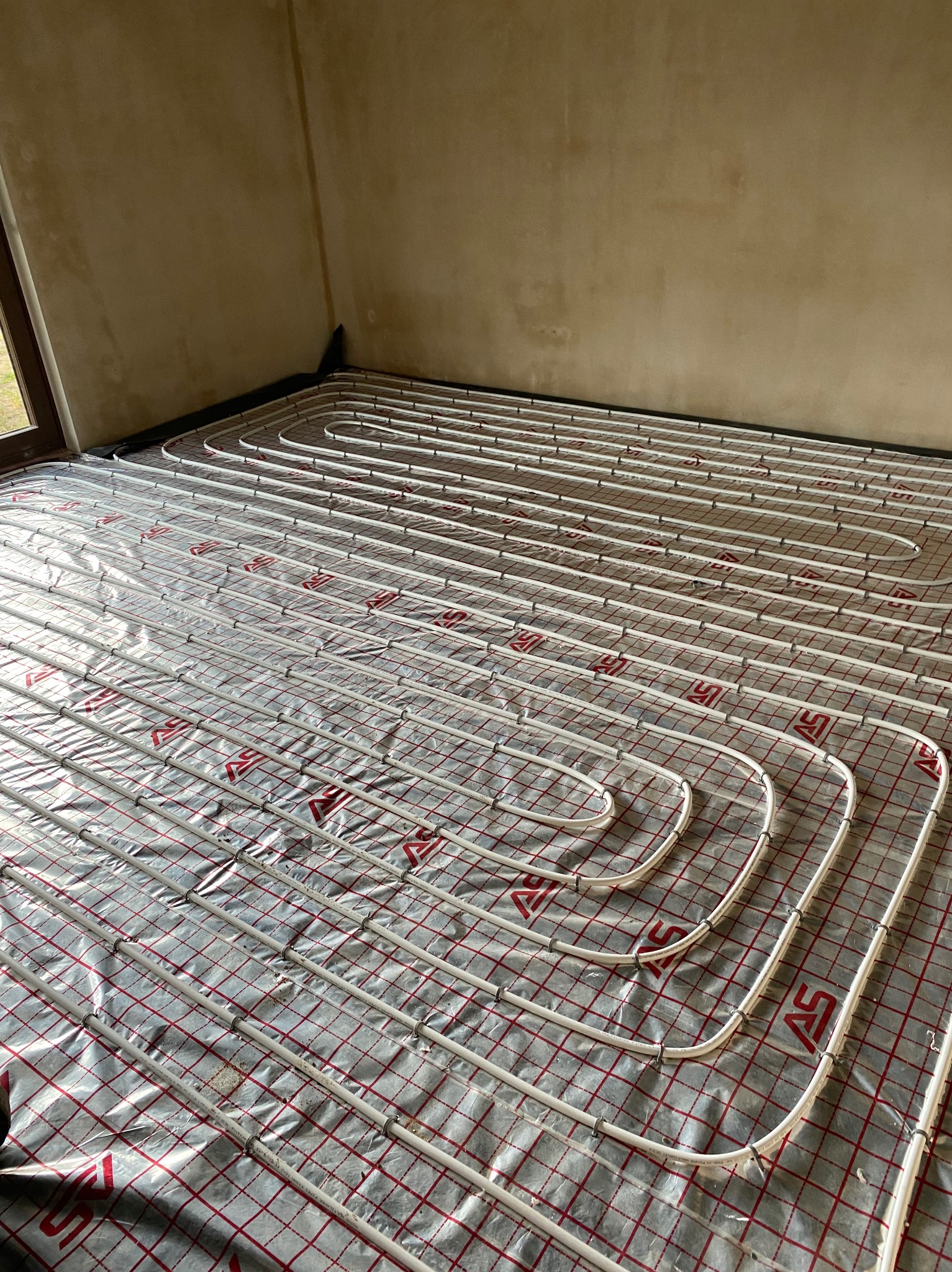 Białe rury ogrzewania podłogowego ułożone spiralnie na srebrnej folii z czerwoną kratką, widok z góry, surowy stan ścian w tle.