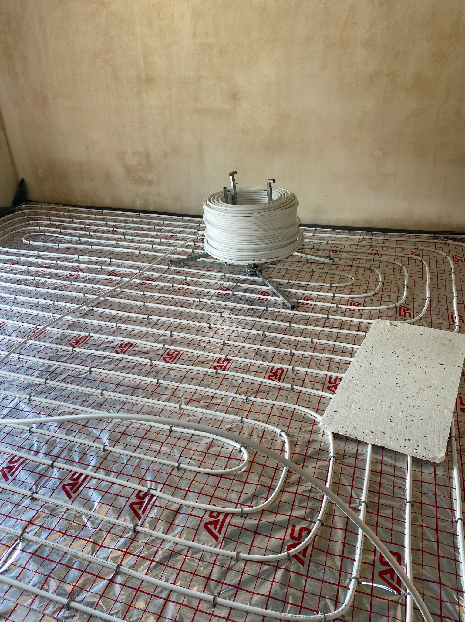 Białe rury ogrzewania podłogowego rozłożone na folii z czerwoną kratką, widoczny zwój rur na stojaku i płyta styropianowa, surowa ściana w tle.