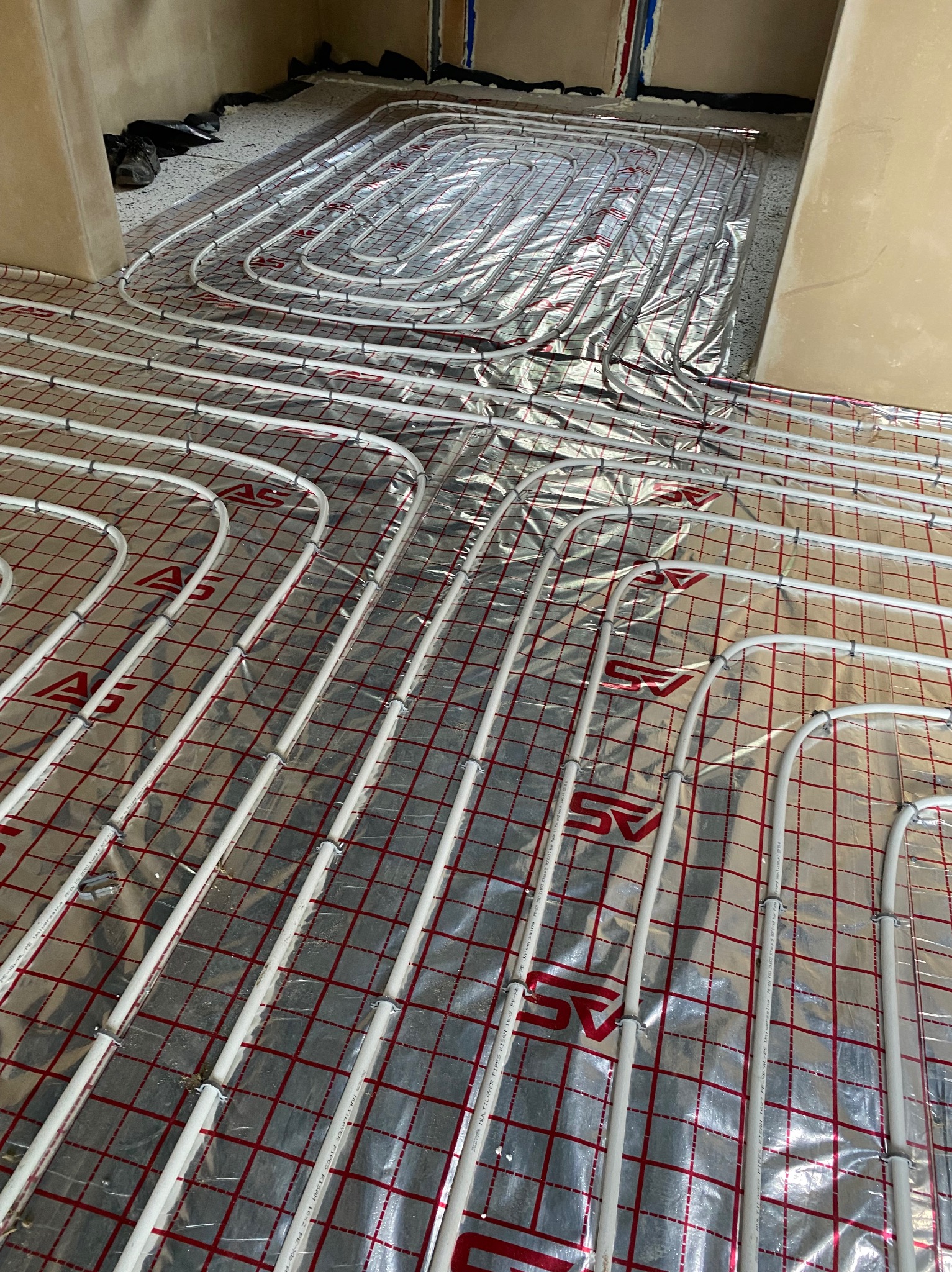 Rozłożone białe rury systemu ogrzewania podłogowego na folii termoizolacyjnej z czerwoną siatką, widoczne złącza i napisy na rurach, surowy stan wykończenia pomieszczenia.