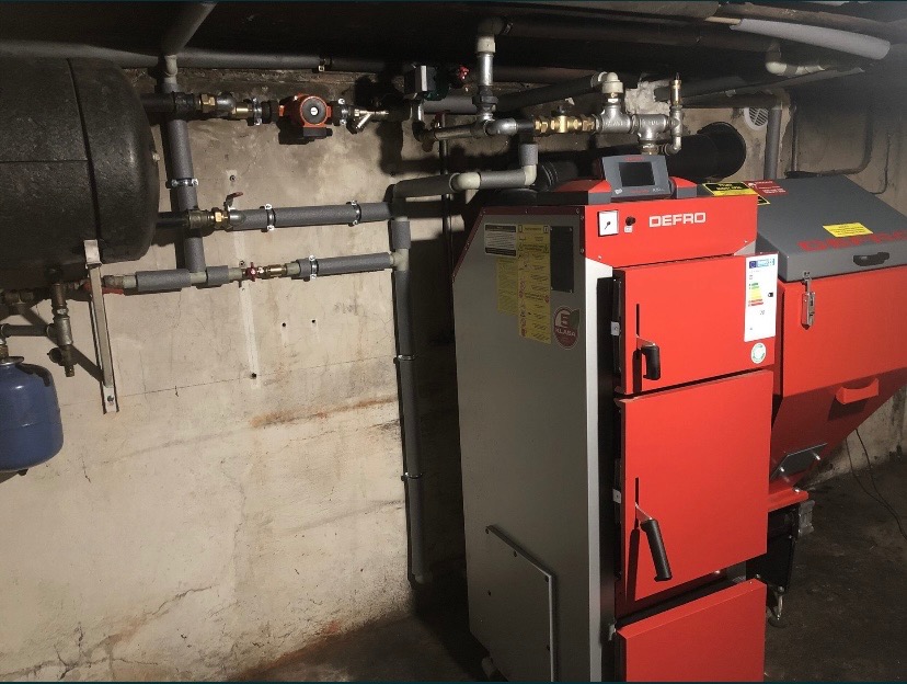 Czerwono-szary piec marki Defro z podajnikiem, zainstalowany w piwnicy z widocznymi rurami instalacji grzewczej i zbiornikiem wyrównawczym.