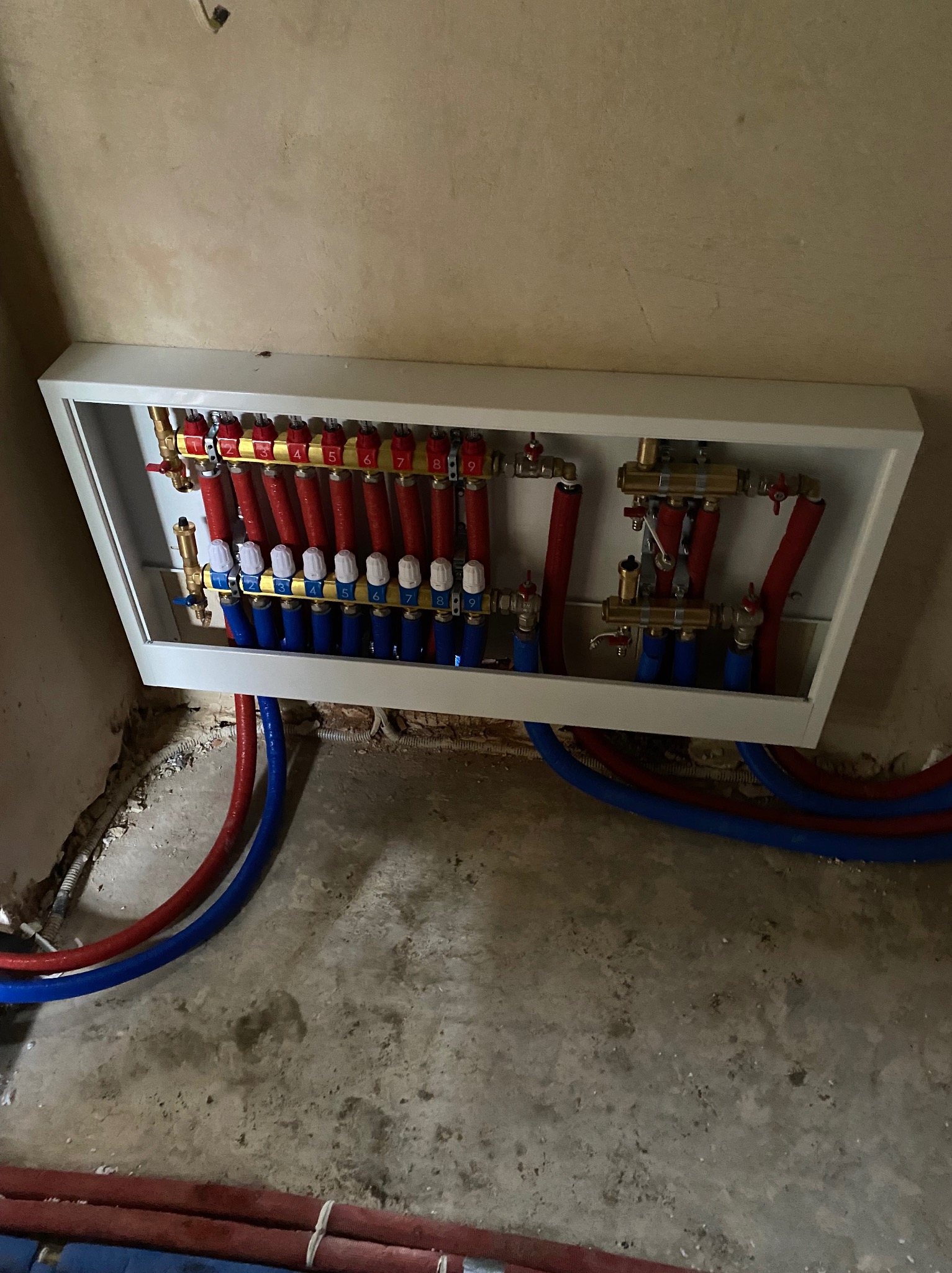 Rozdzielacz ogrzewania podłogowego w białej obudowie, z czerwonymi i niebieskimi rurami, numery na górze, widoczne złącza i zawory, surowy stan podłogi i ściany.