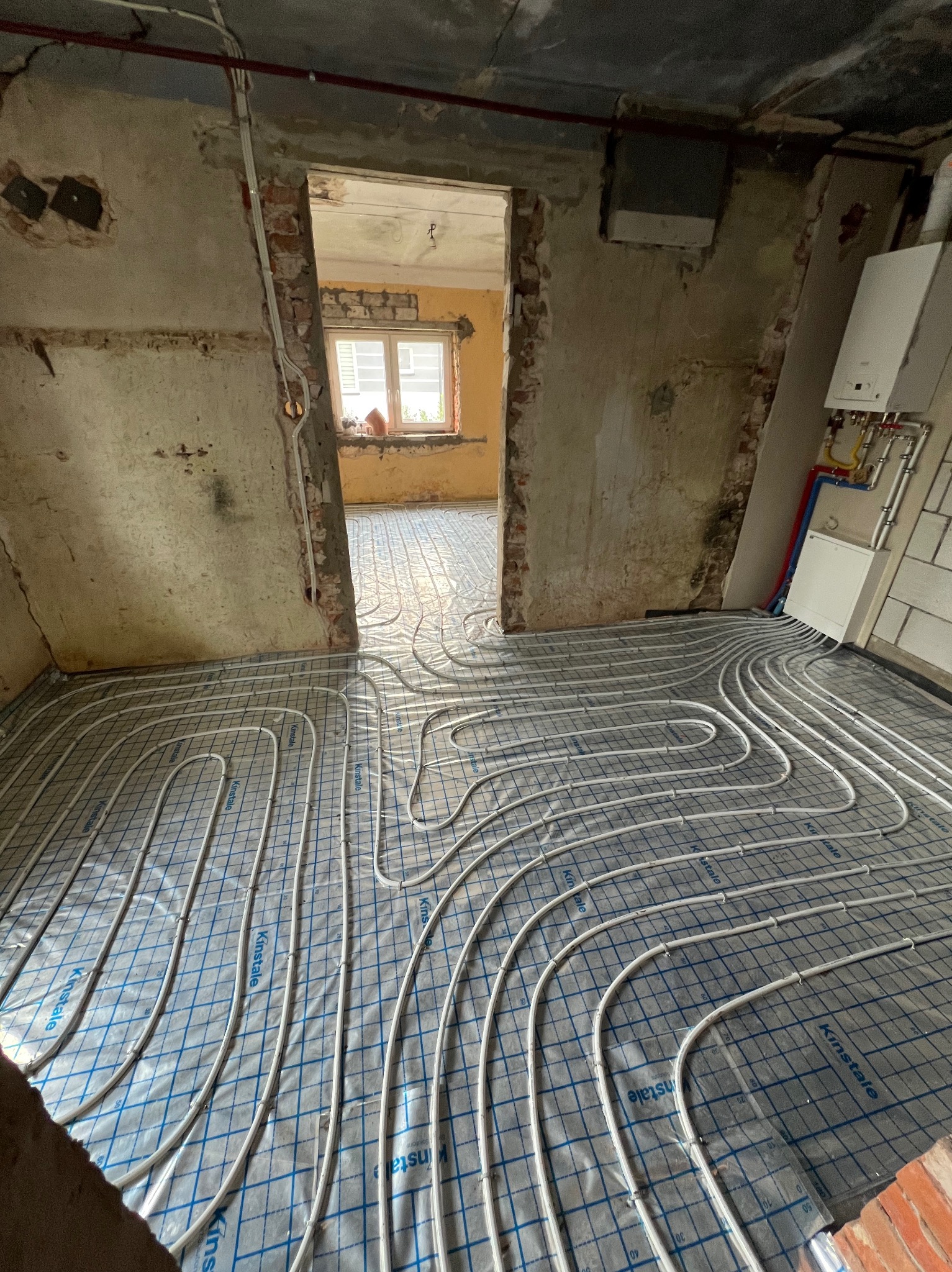 Instalacja ogrzewania podłogowego w surowym wnętrzu: białe rury ułożone na folii z nadrukiem Kinstale, ściany w stanie deweloperskim, widoczny piec gazowy w tle.