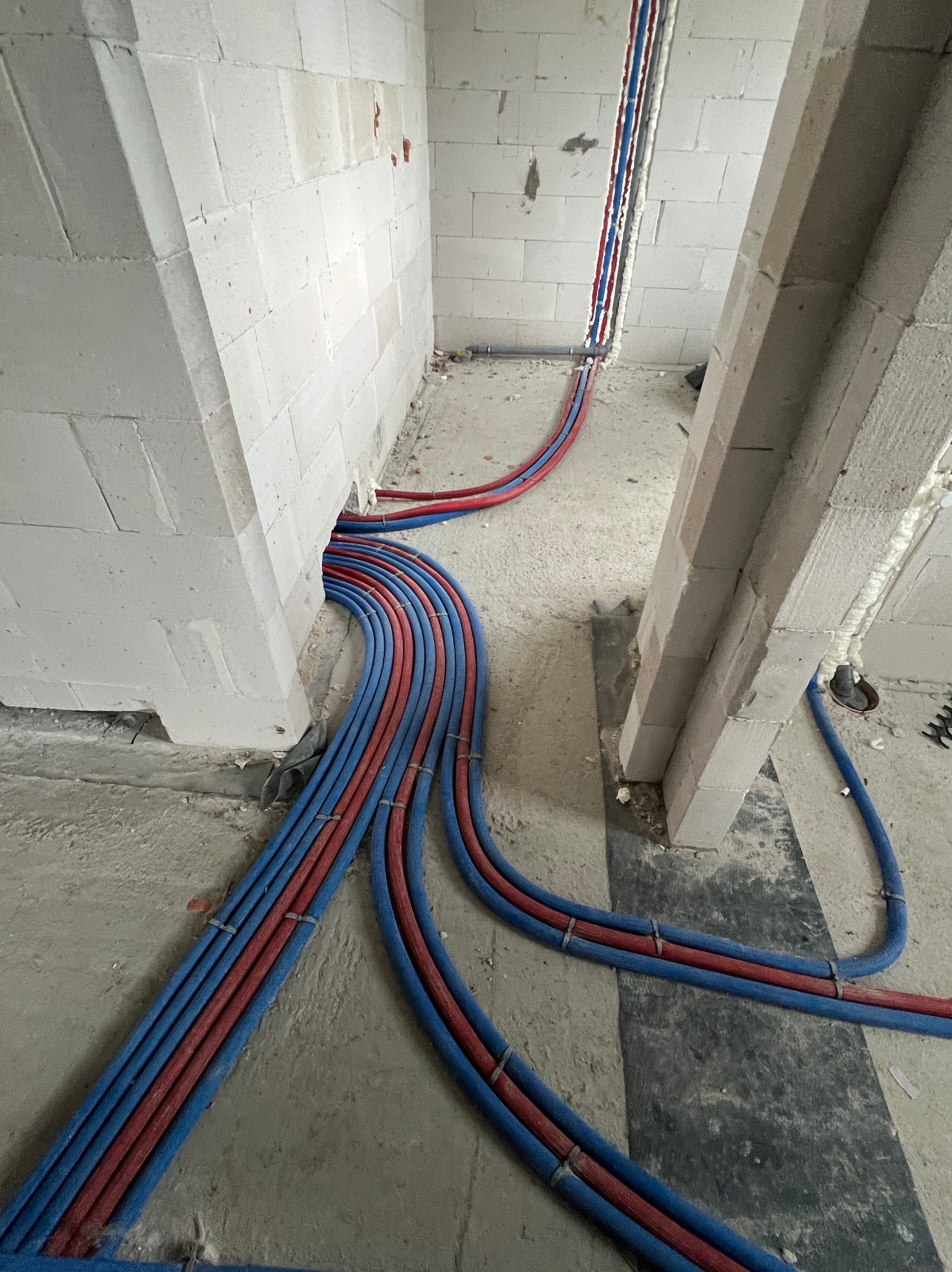 Instalacja ogrzewania podłogowego: czerwono-niebieskie rury prowadzone po surowym podłożu między ścianami z betonu komórkowego, widoczne elementy mocujące.