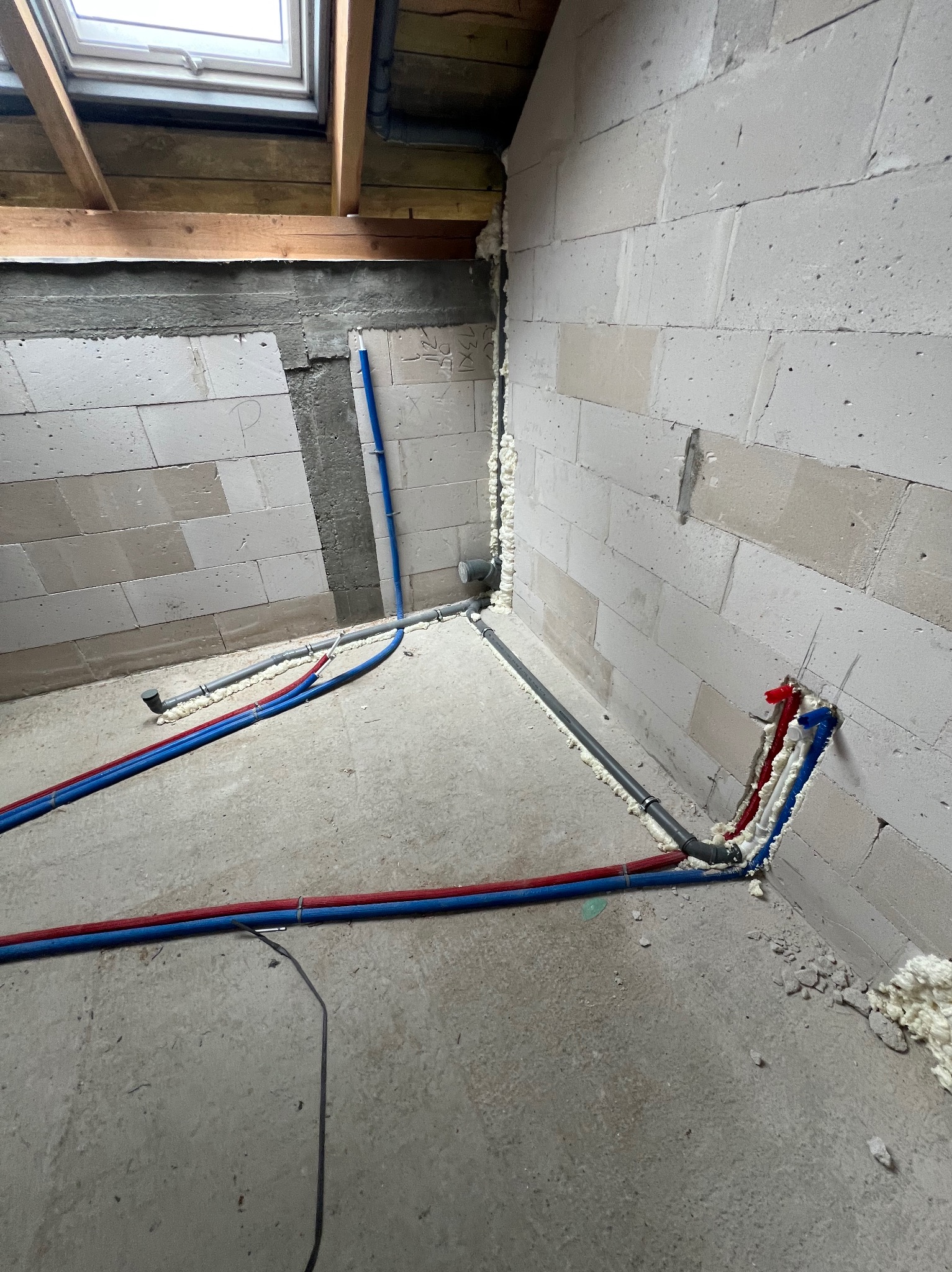 Wnętrze w trakcie prac hydraulicznych: widoczne rury (czerwone, niebieskie, szare) prowadzone wzdłuż ścian z betonu komórkowego. Okno dachowe w tle.