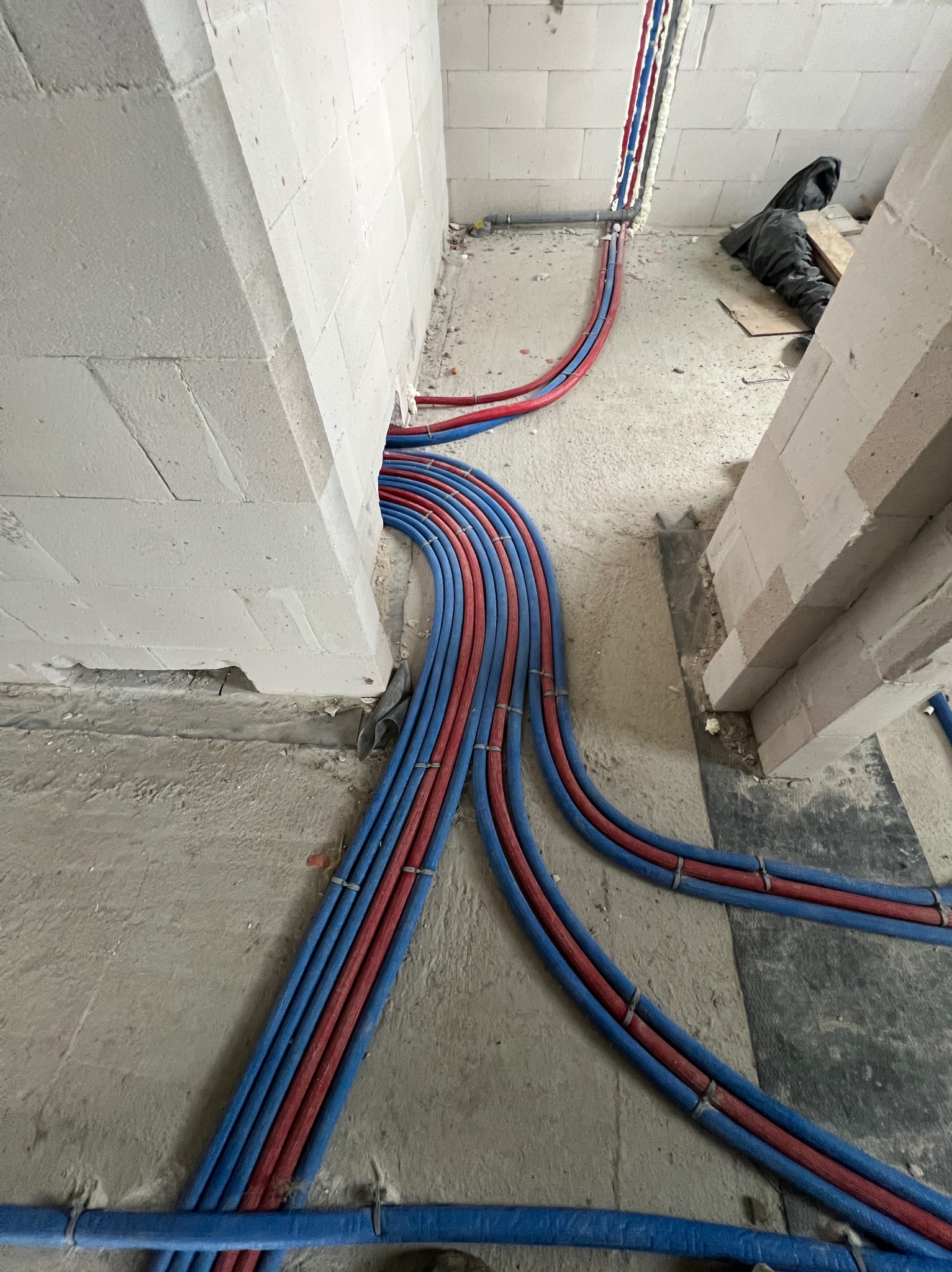 Instalacja ogrzewania podłogowego w surowym wnętrzu. Ułożone rury grzewcze w kolorach niebieskim i czerwonym, przymocowane do podłoża. Widoczne ściany z bloczków.