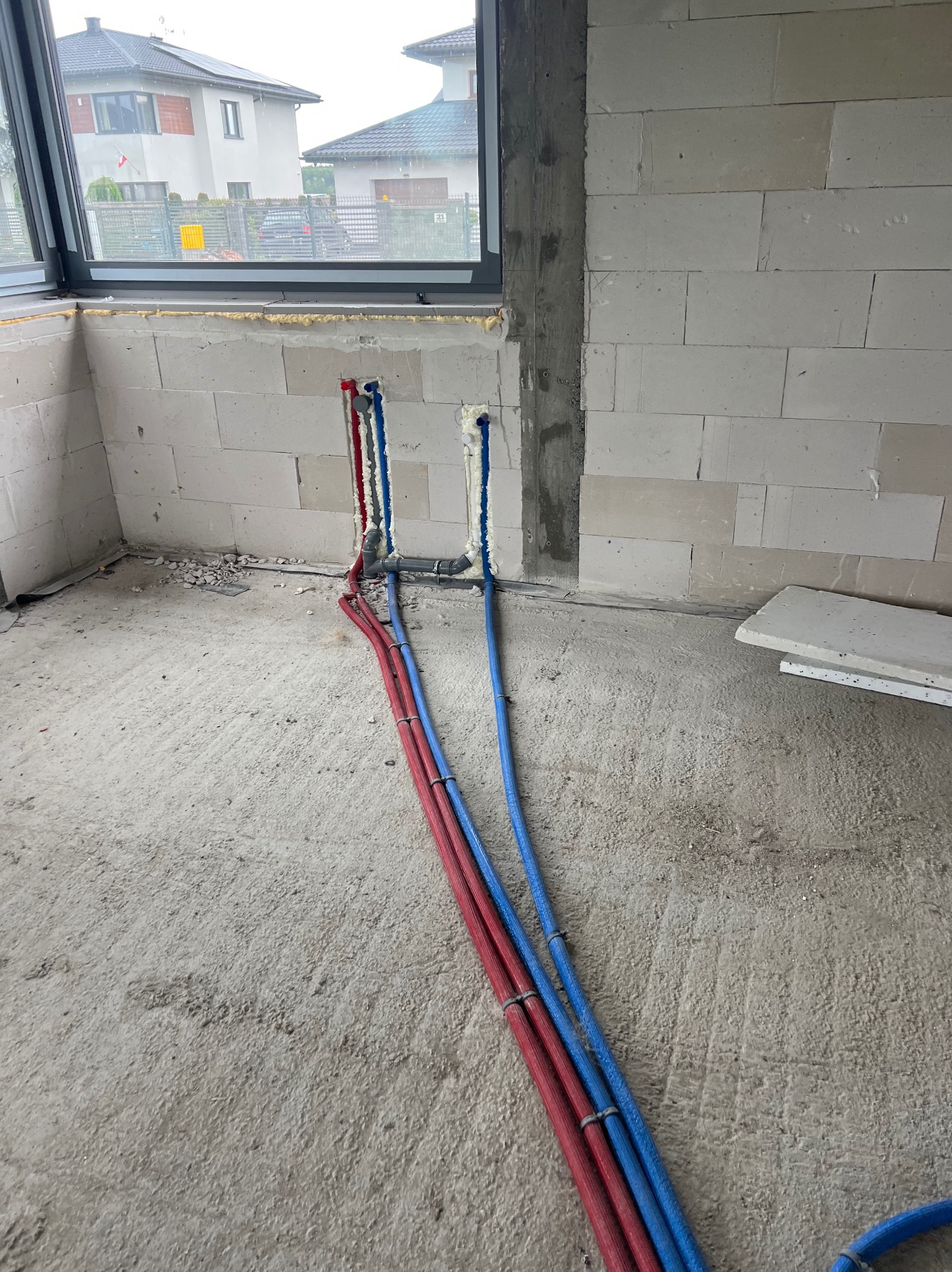 Wnętrze w trakcie prac hydraulicznych: czerwone i niebieskie rury prowadzone wzdłuż ściany z betonu komórkowego, widoczne elementy instalacji przy oknie.