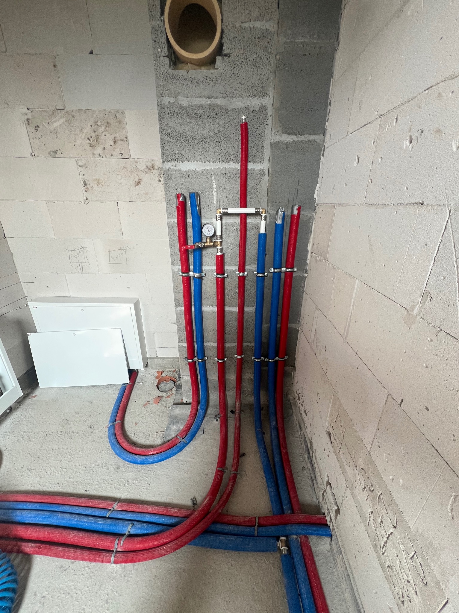 Instalacja wodna z rur PEX w kolorze niebieskim i czerwonym, zamontowana przy szarej ścianie z betonu komórkowego. Manometr i zawory przy pionowych rurach.