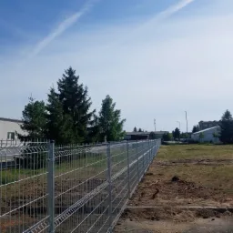 Długa, metalowa siatka ogrodzeniowa, widok z boku, z zielonymi drzewami i budynkami w tle, na ziemi widoczne ślady po pracach ziemnych.