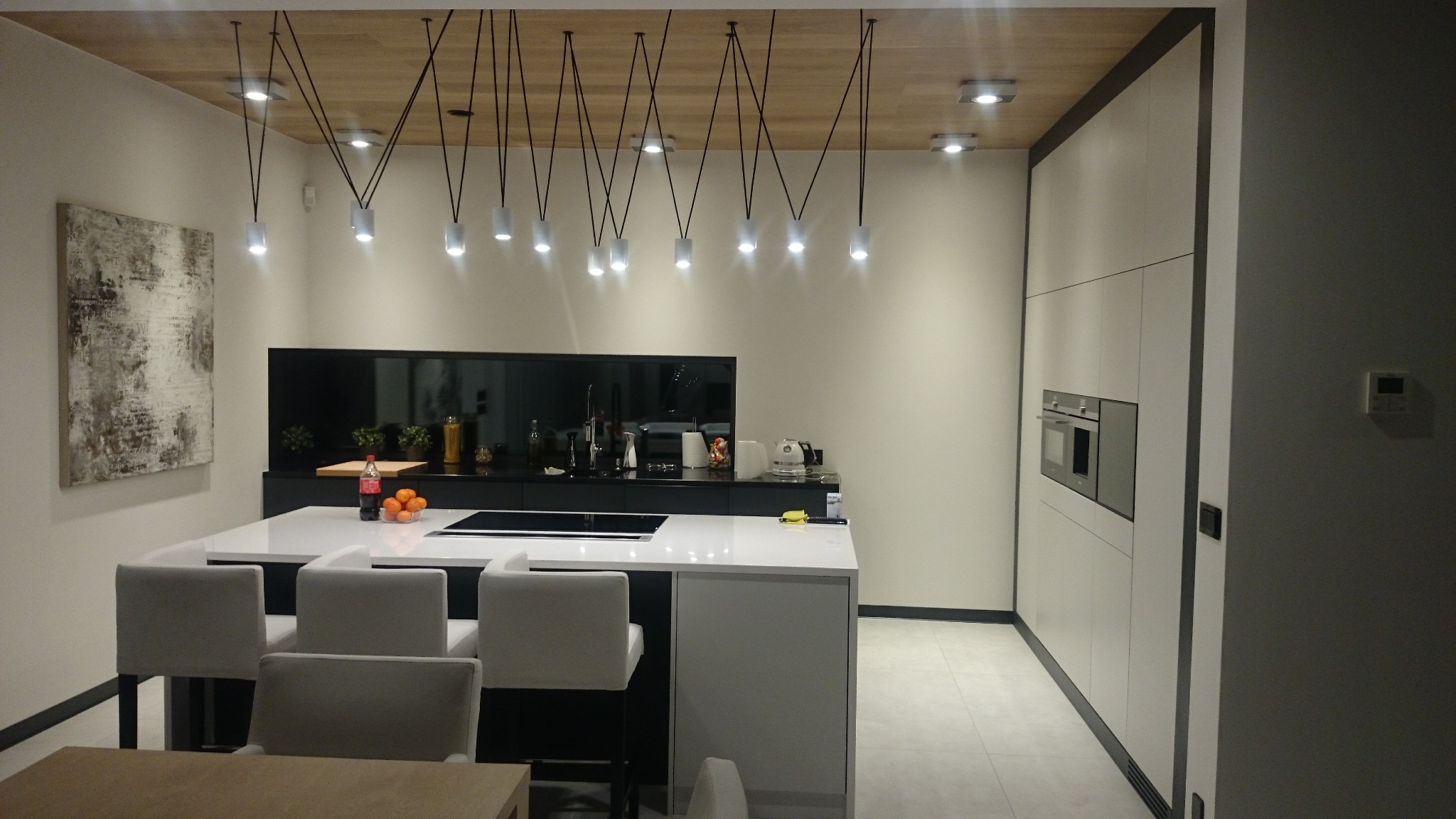 Nowoczesna kuchnia z wyspą, białymi szafkami i oryginalnym oświetleniem punktowym na drewnianym suficie, z minimalistycznym obrazem na ścianie.