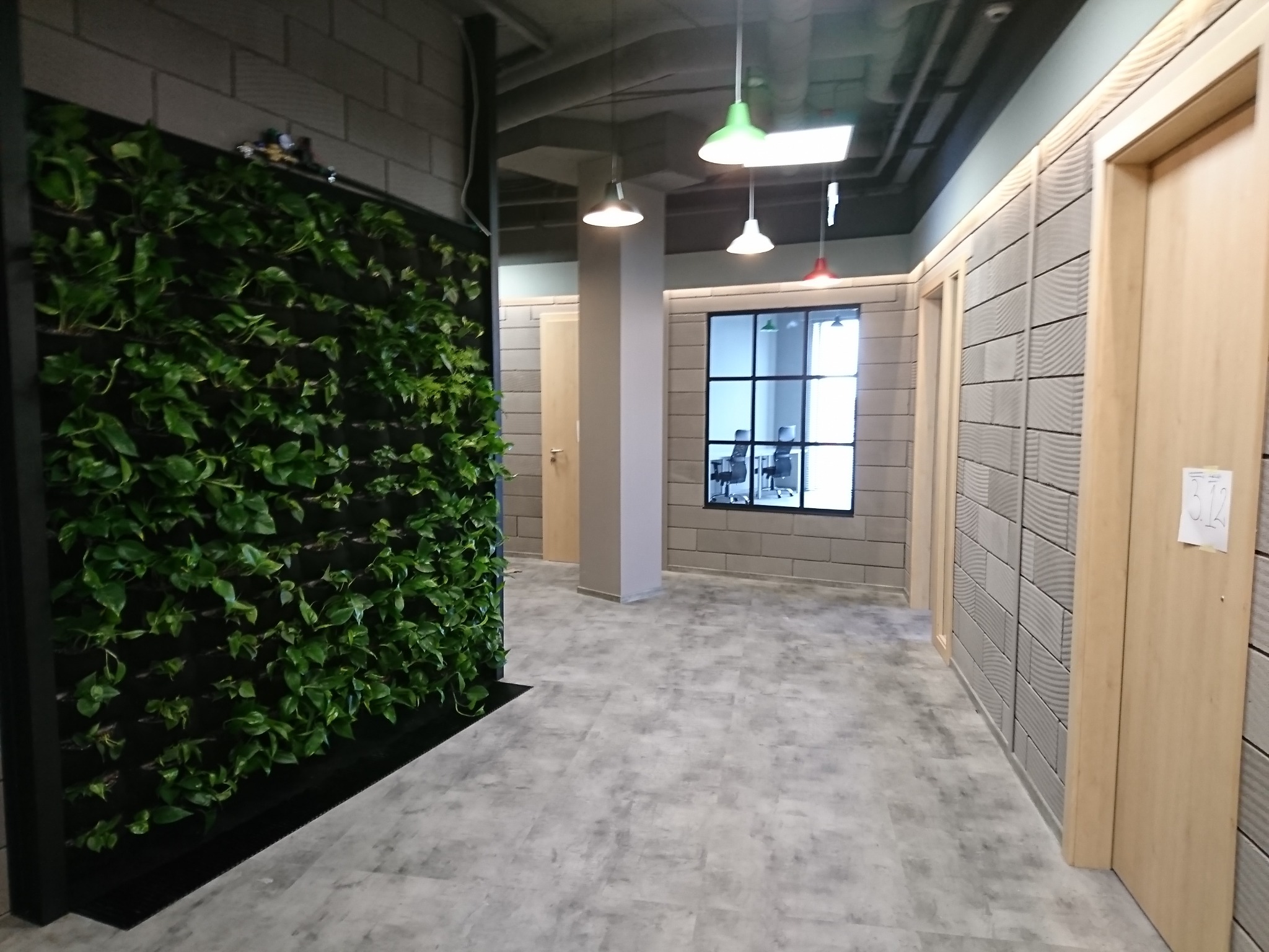 Wnętrze biura z pionową ścianą zieleni, szarą podłogą i ścianami z dekoracyjnymi panelami. Widoczne wiszące lampy z kolorowymi kloszami.