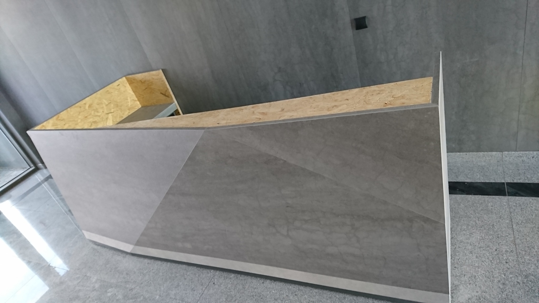 Wnętrze biura w trakcie wykańczania, widoczna surowa konstrukcja lady recepcyjnej z płyty OSB i szarej okładziny imitującej kamień, na podłodze płytki w odcieniach szarości.