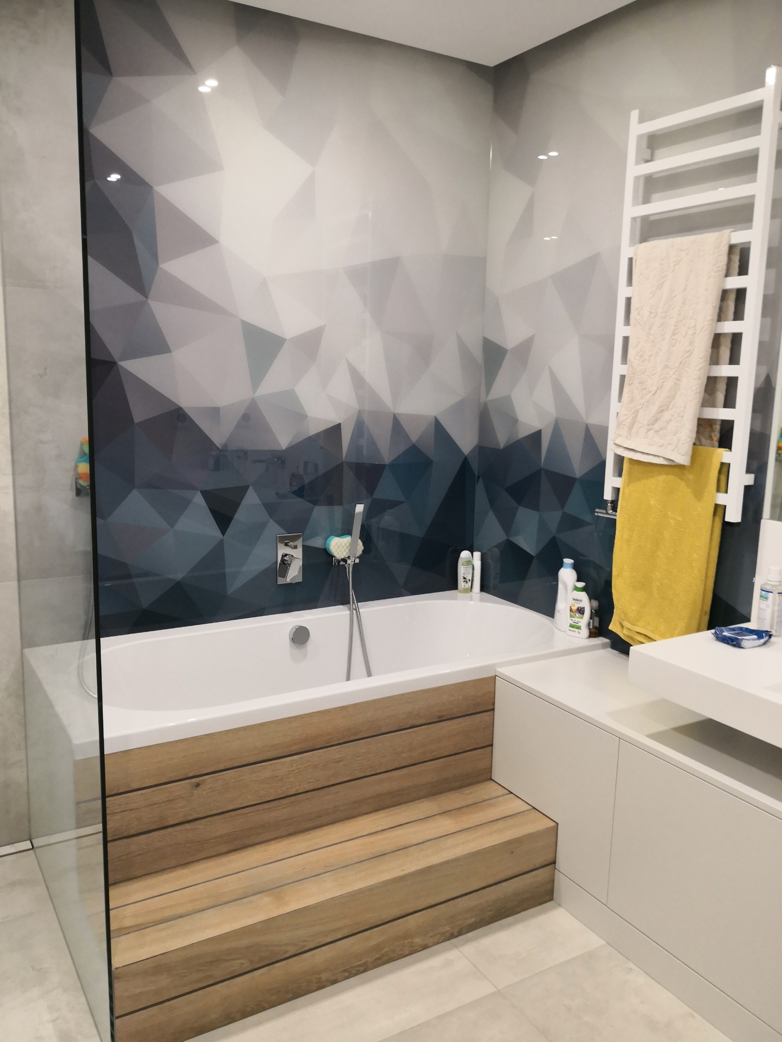 Nowoczesna łazienka w Warszawie z wanną obudowaną drewnopodobnymi panelami, geometryczną tapetą w odcieniach szarości i błękitu oraz grzejnikiem drabinkowym z beżowym i żółtym ręcznikiem.