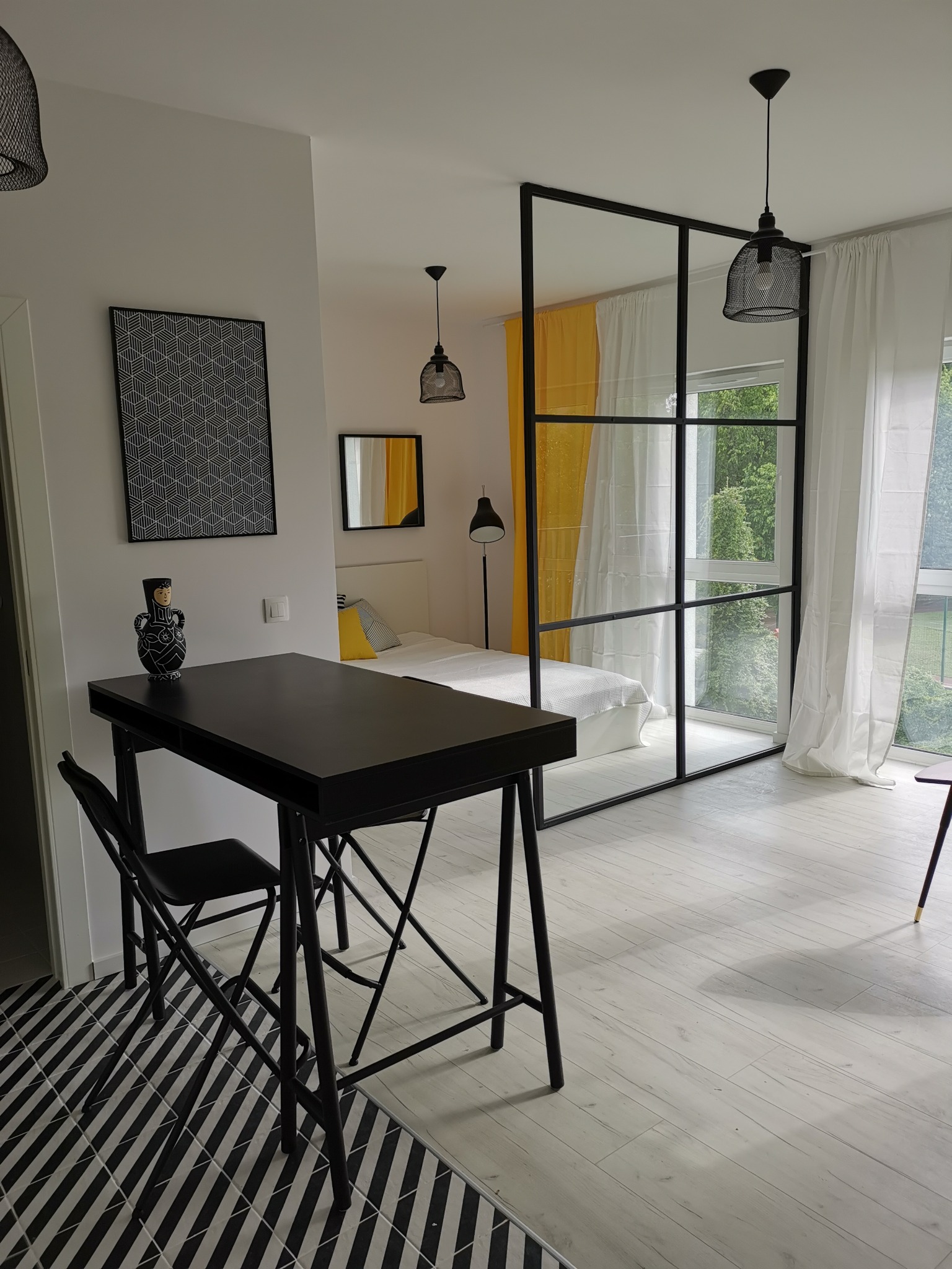 Nowoczesne wnętrze mieszkania z czarnym stołem i krzesłem na tle sypialni oddzielonej szklaną ścianką z czarnymi ramami, widok na łóżko z żółtą poduszką i oknem z białymi zasłonami.