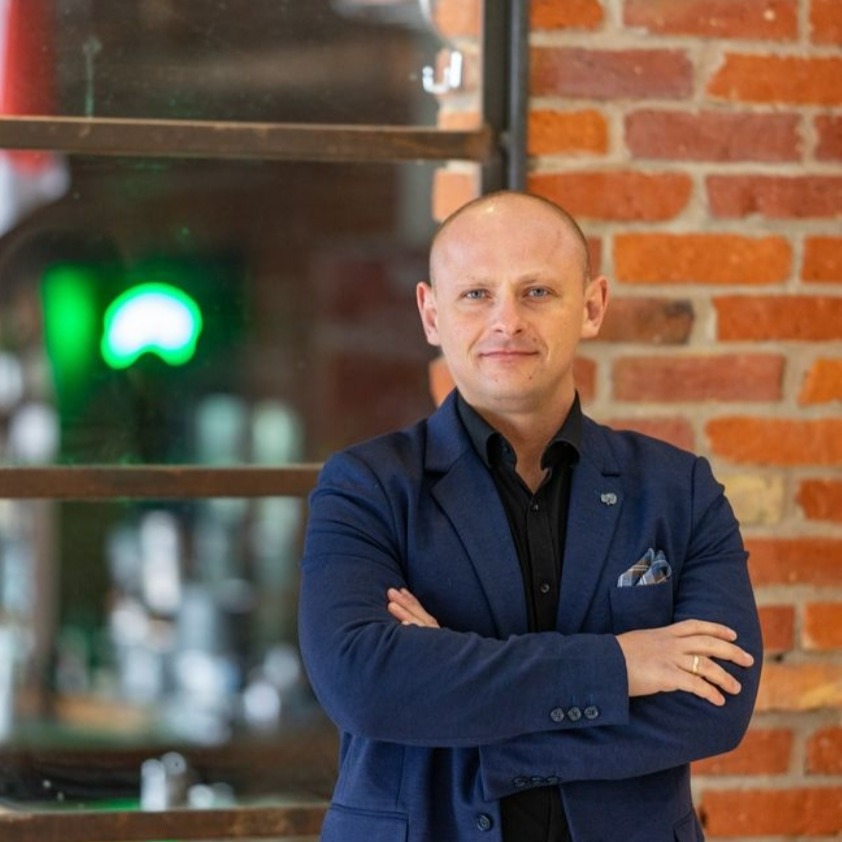 Uśmiechnięty mężczyzna w niebieskiej marynarce i czarnej koszuli, stojący na tle ceglanej ściany i okna z zielonym neonem.