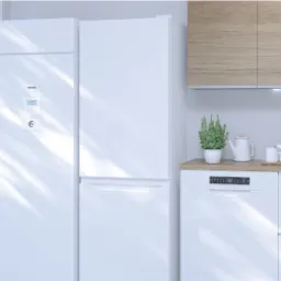 Biała pompa ciepła Panasonic zainstalowana na balkonie mieszkania z widokiem na miasto, obok zabudowy kuchennej z lodówką i zmywarką.