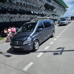Konrad Bek TOURS STELABUS - Szary van Mercedes parkuje przed nowoczesnym budynkiem z lustrzaną fasadą. W tle widoczni ludzie i inne pojazdy. Słoneczny dzień, błękitne niebo.