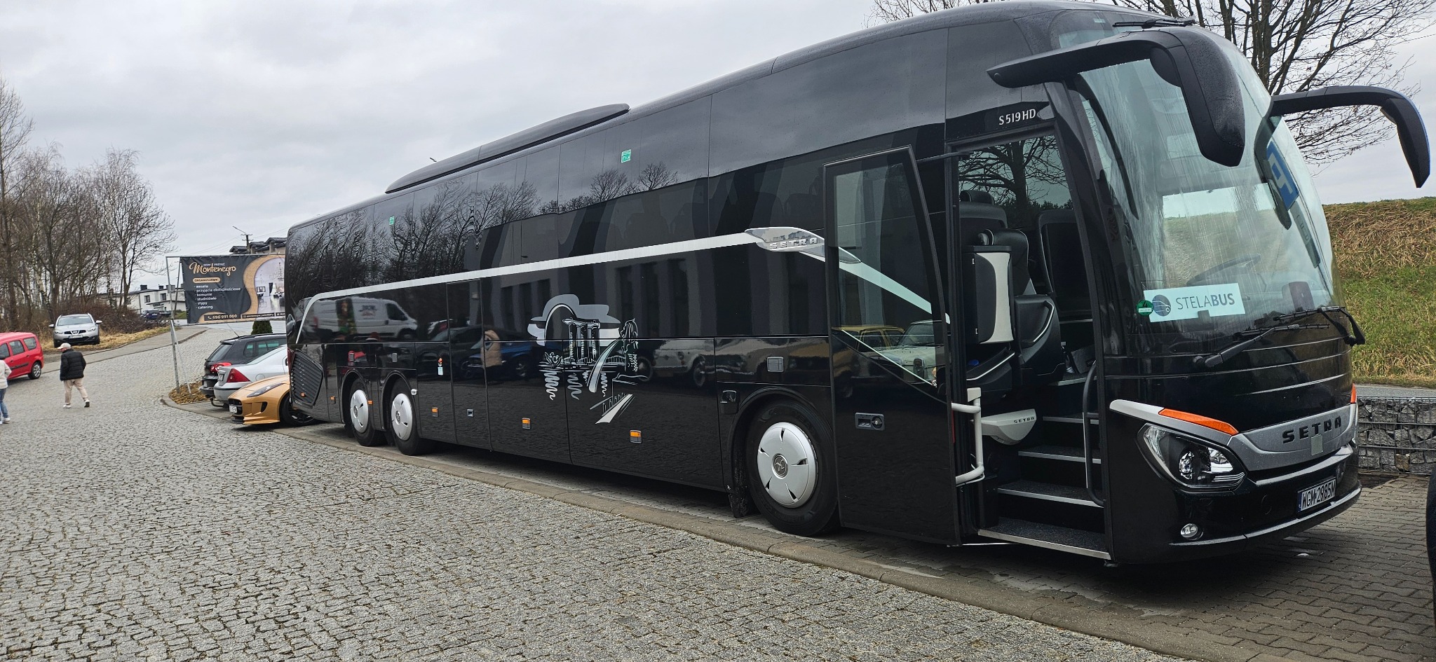 Czarny autokar Setra S519HD z otwartymi drzwiami, zaparkowany na brukowanej powierzchni, z logo Stelabus na przedniej szybie i grafiką z motywem miejskim na boku.