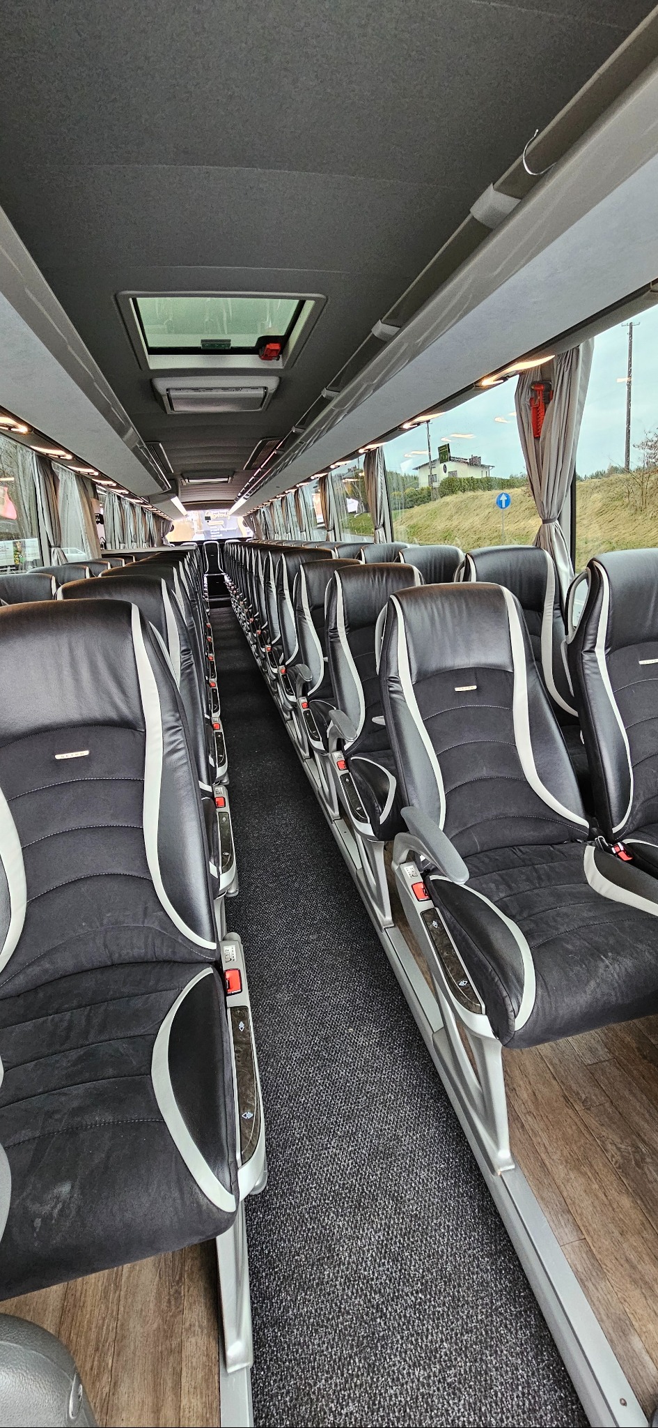 Wnętrze autobusu turystycznego z rzędami czarno-białych skórzanych foteli, widok na okno z krajobrazem i znak drogowy.