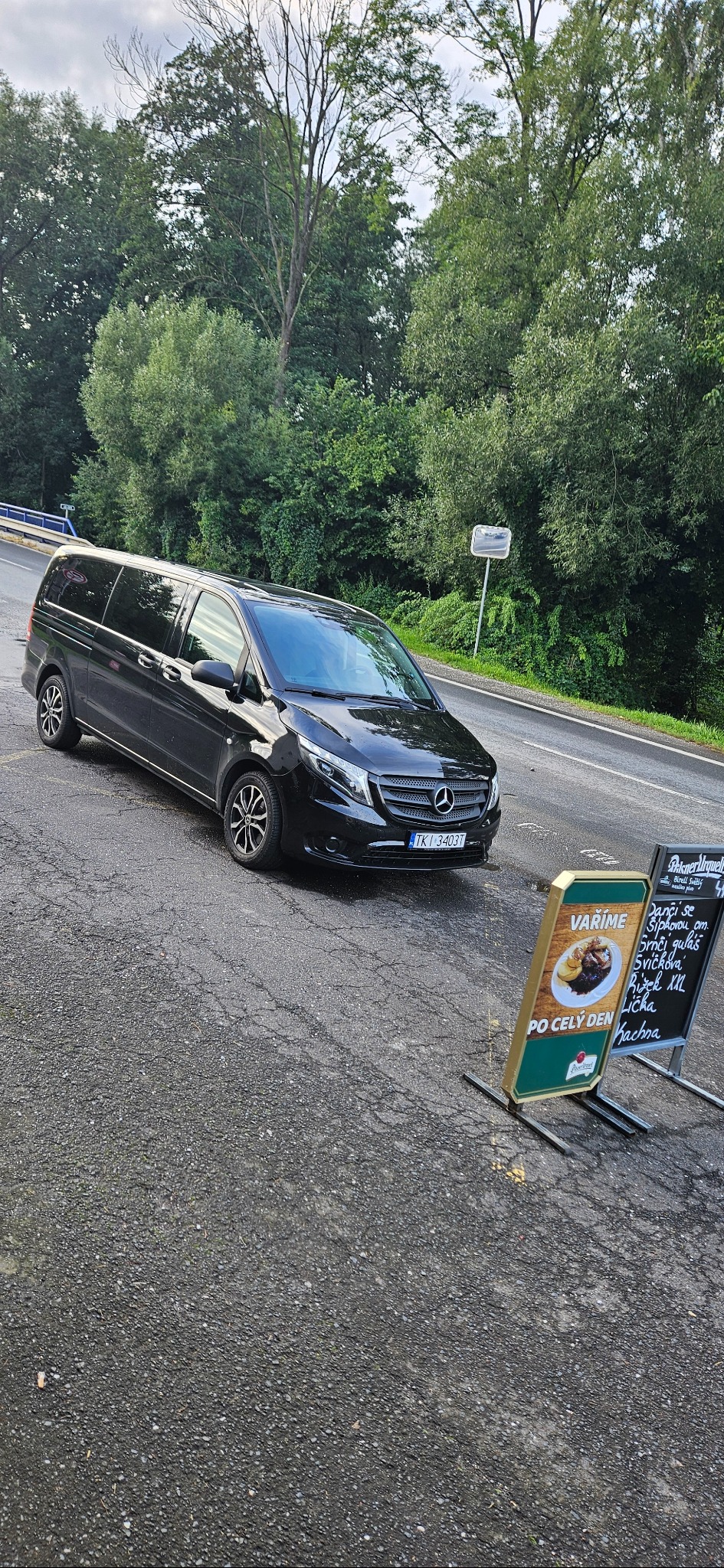 Czarny van Mercedes V-klasy z numerem TKJ 3403T zaparkowany na poboczu drogi, obok tablic reklamowych restauracji z menu w języku czeskim.