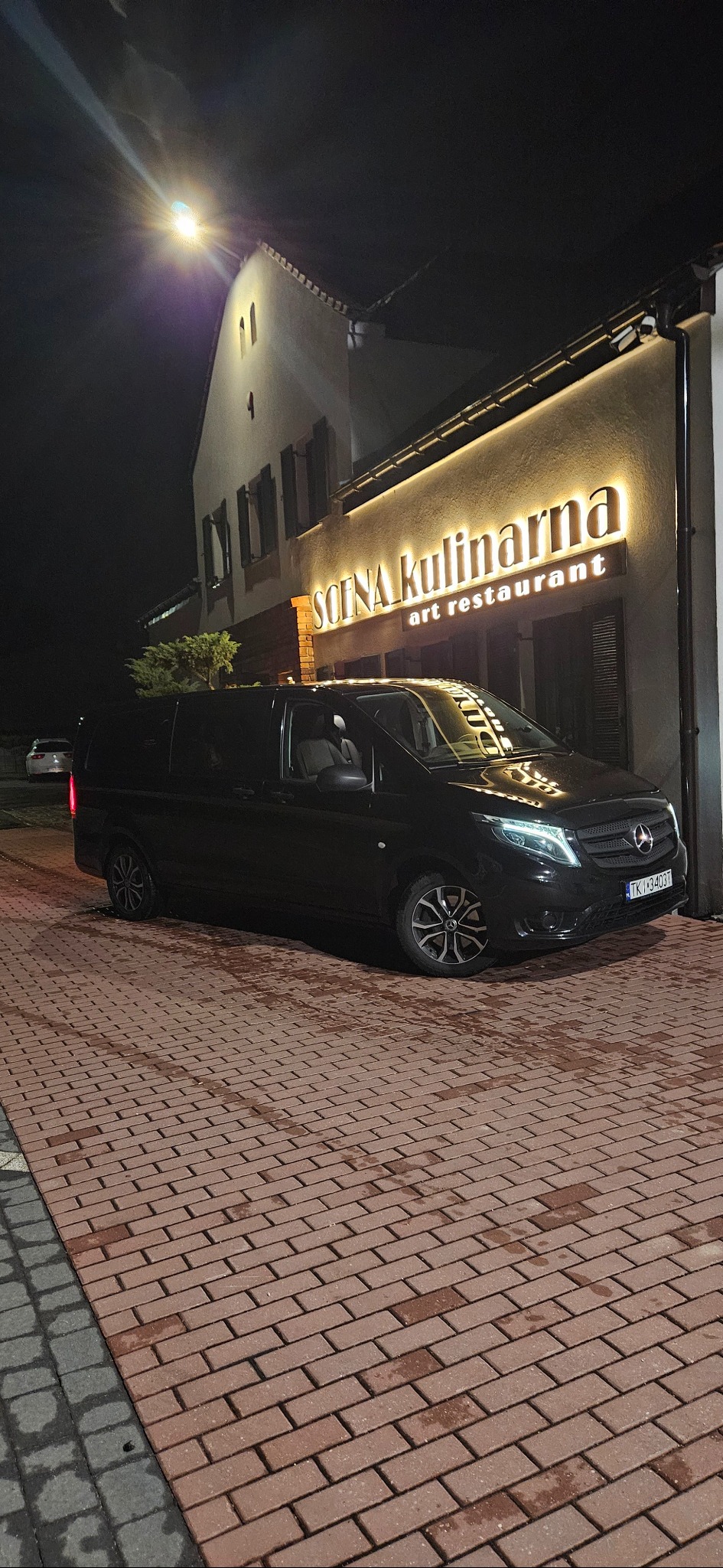 Czarny van Mercedes-Benz V-klasy zaparkowany nocą przed restauracją z podświetlanym szyldem, widoczne detale felg i reflektorów. Czerwona kostka brukowa na parkingu.