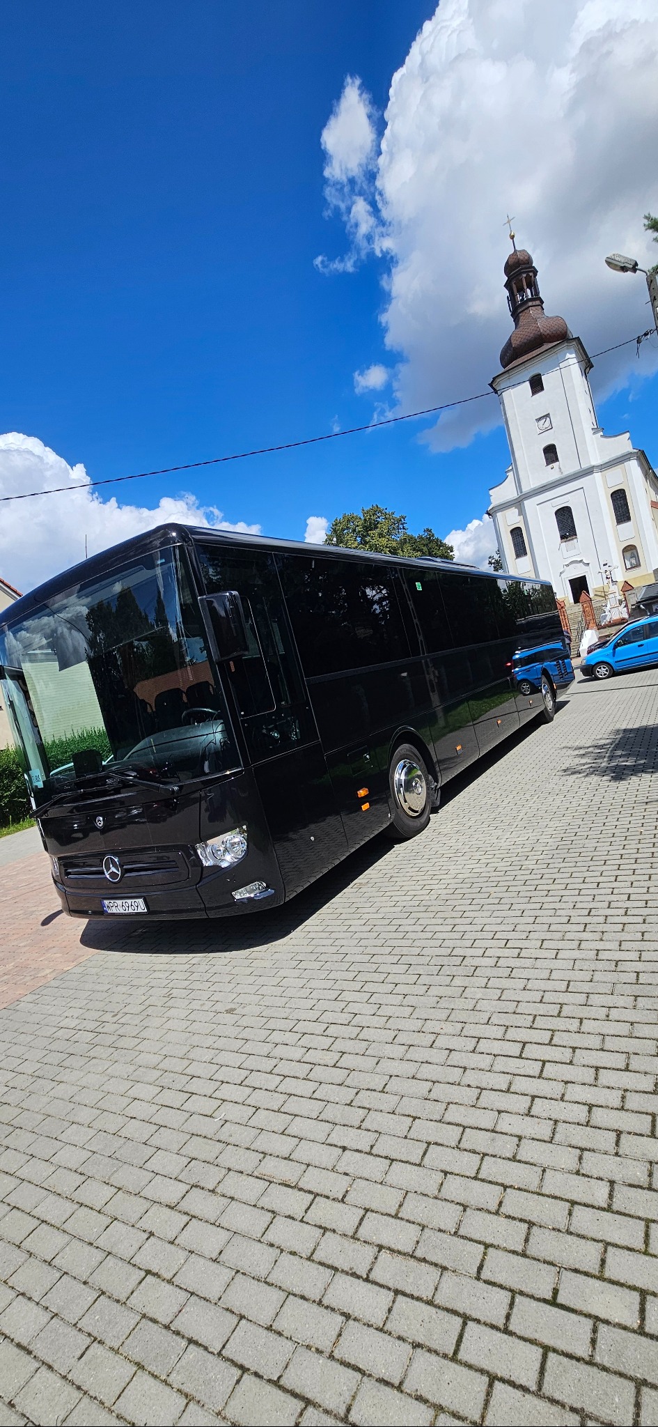 Czarny autokar Mercedes na brukowanej ulicy, z kościołem w tle pod błękitnym niebem. Widoczny numer rejestracyjny i logo Mercedes-Benz.