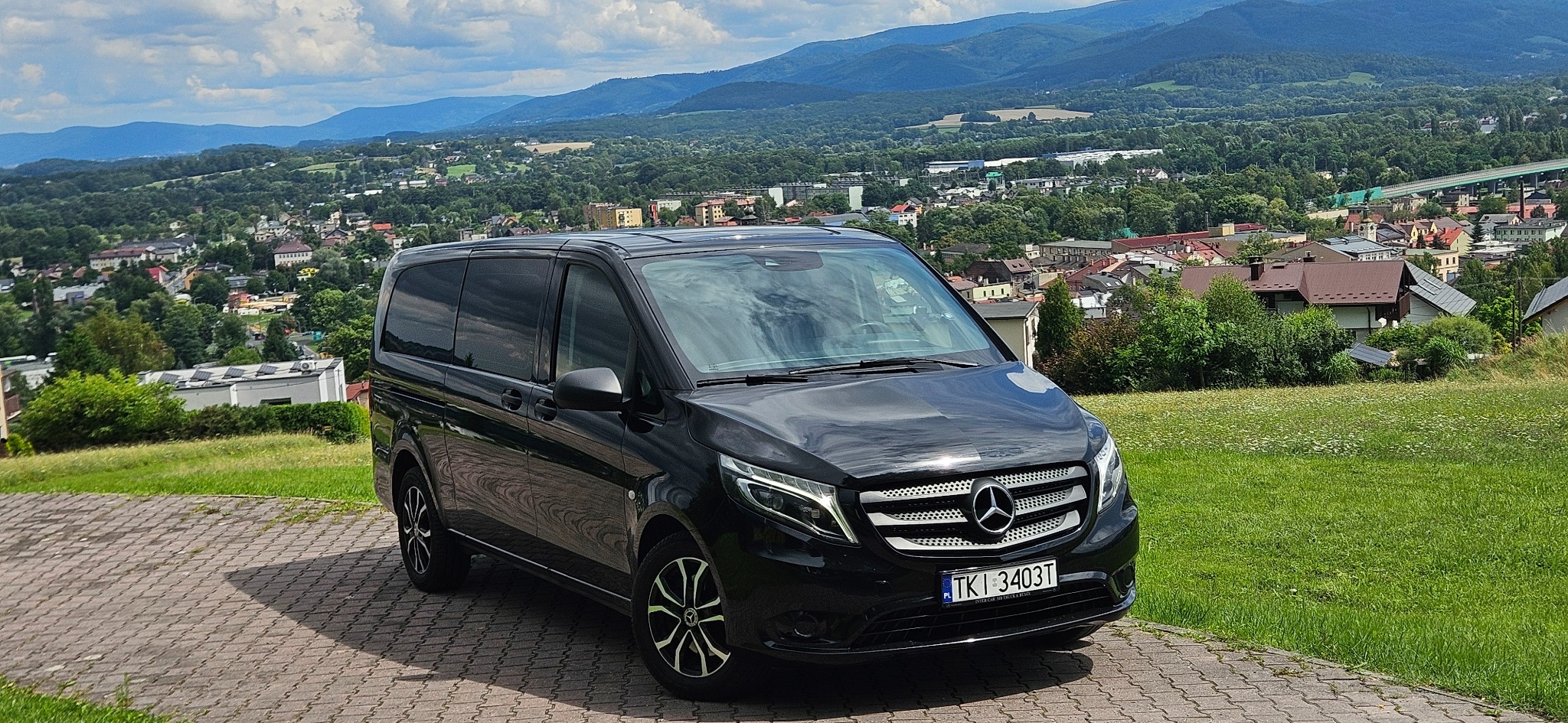 Czarny van Mercedes-Benz z polską rejestracją na tle malowniczego krajobrazu górskiego i zabudowy miejskiej. Auto zaparkowane na brukowanej nawierzchni.