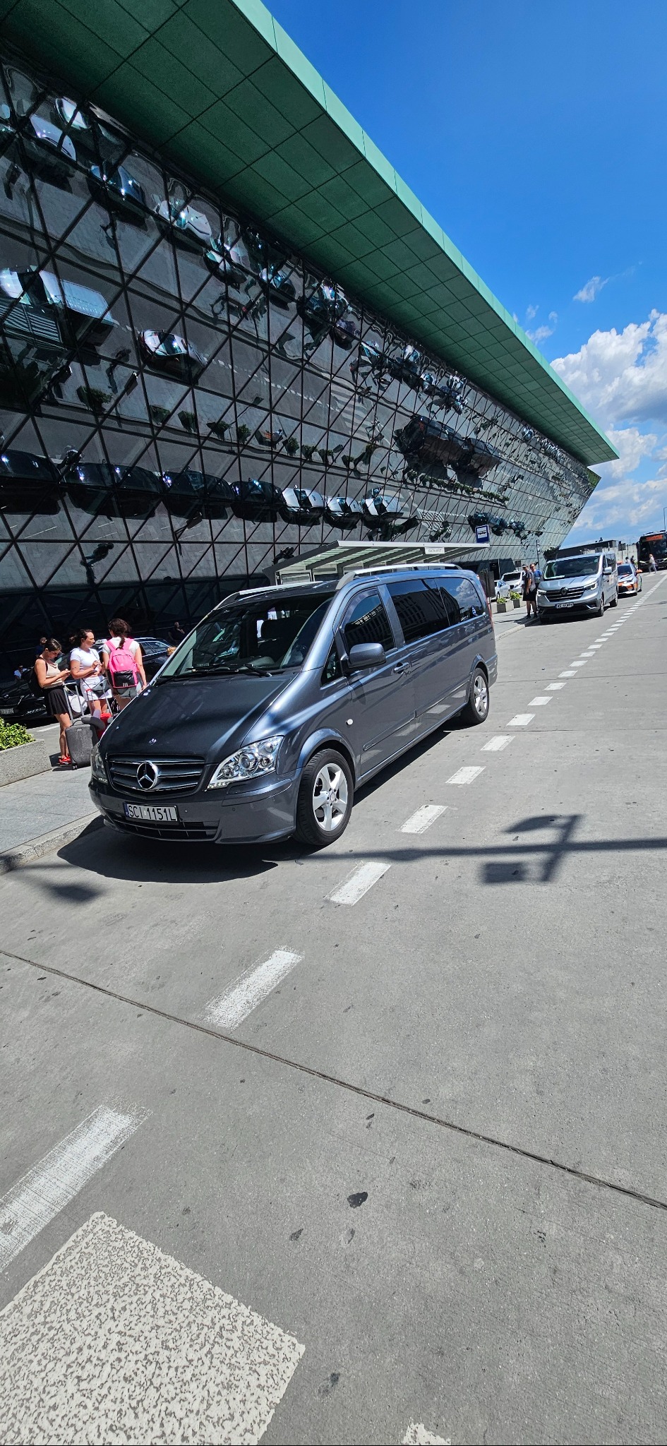 Szary van Mercedes parkuje przed nowoczesnym budynkiem z lustrzaną fasadą. W tle widoczni ludzie i inne pojazdy. Słoneczny dzień, błękitne niebo.