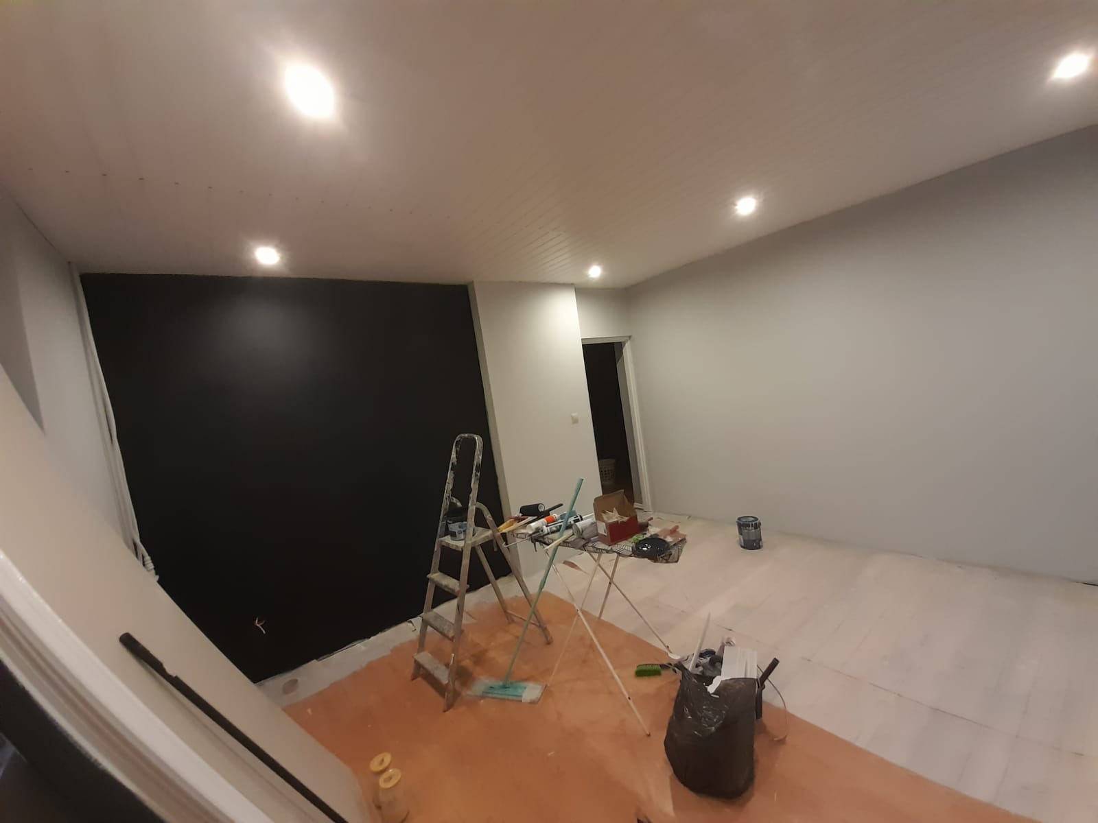 Pokój w trakcie malowania: czarna ściana kontrastuje z białą, na podłodze folia ochronna, drabina i stół z narzędziami malarskimi.