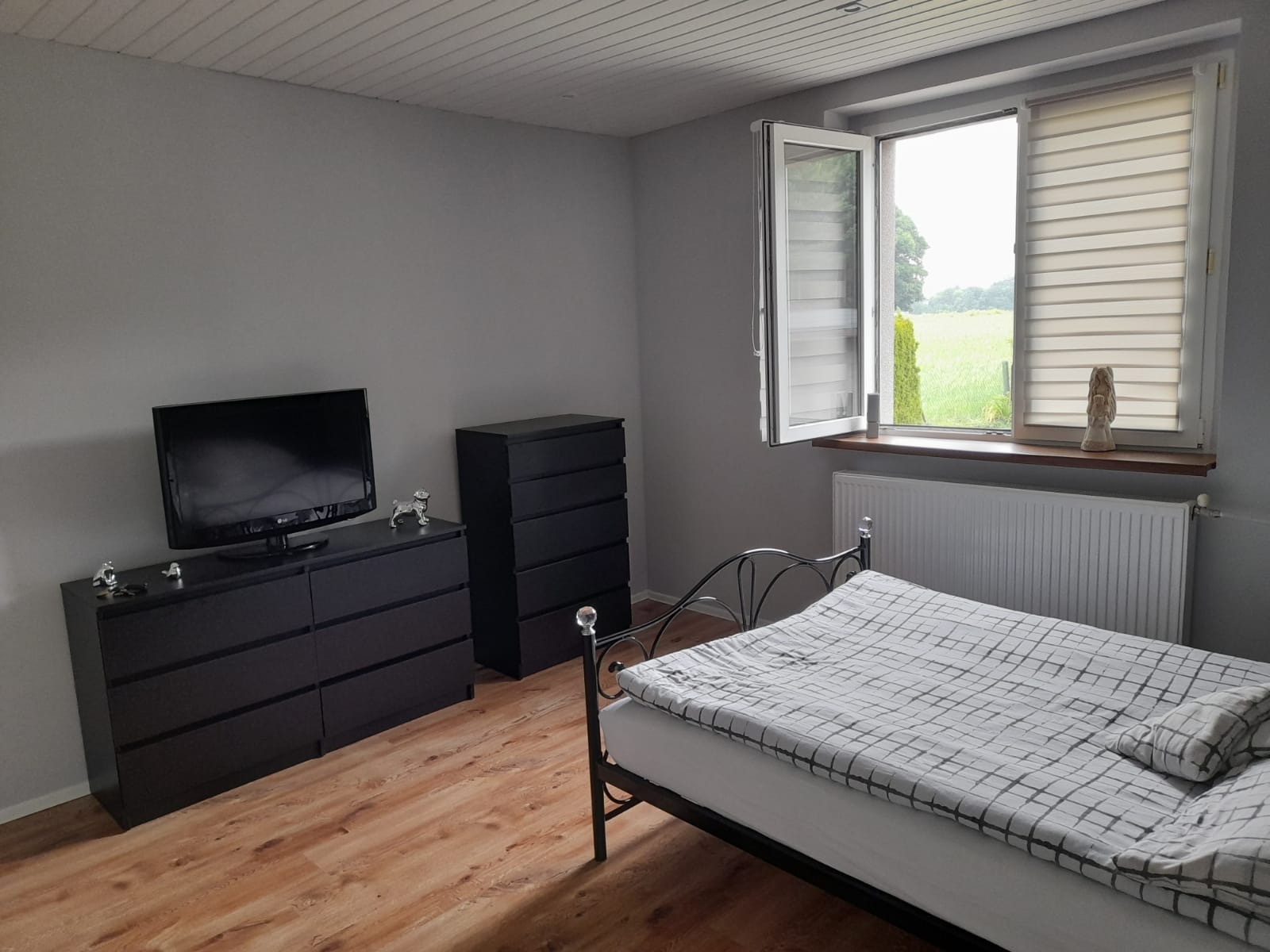 Pokój z szarą ścianą, drewnianą podłogą, czarnymi komodami, telewizorem, łóżkiem z metalową ramą i oknem z widokiem na zieleń.