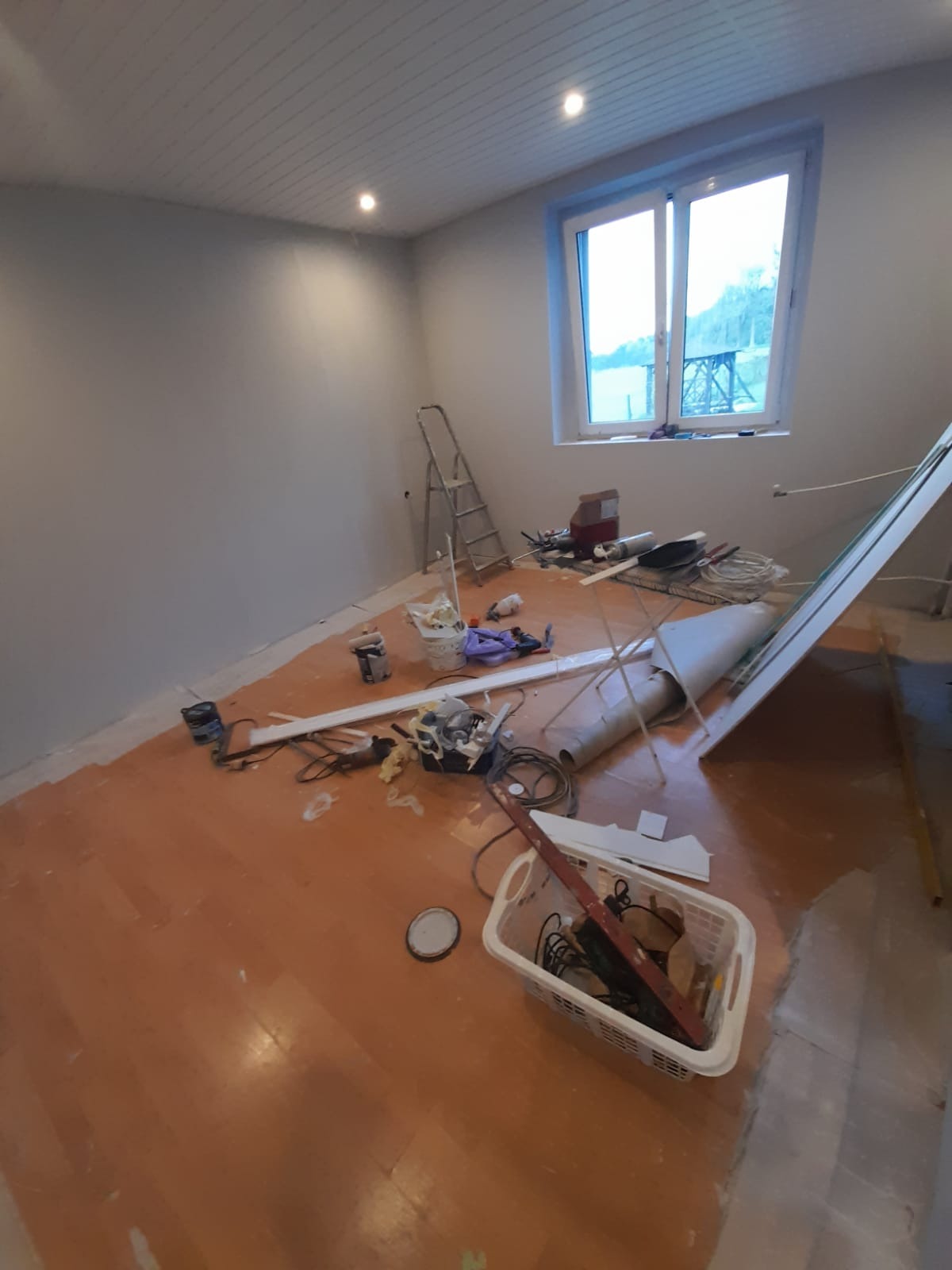 Pokój w trakcie remontu: ściany świeżo pomalowane na szaro, podłoga zabezpieczona, widoczne narzędzia, drabina, okno z widokiem na zewnątrz, plastikowy kosz z narzędziami na pierwszym planie.