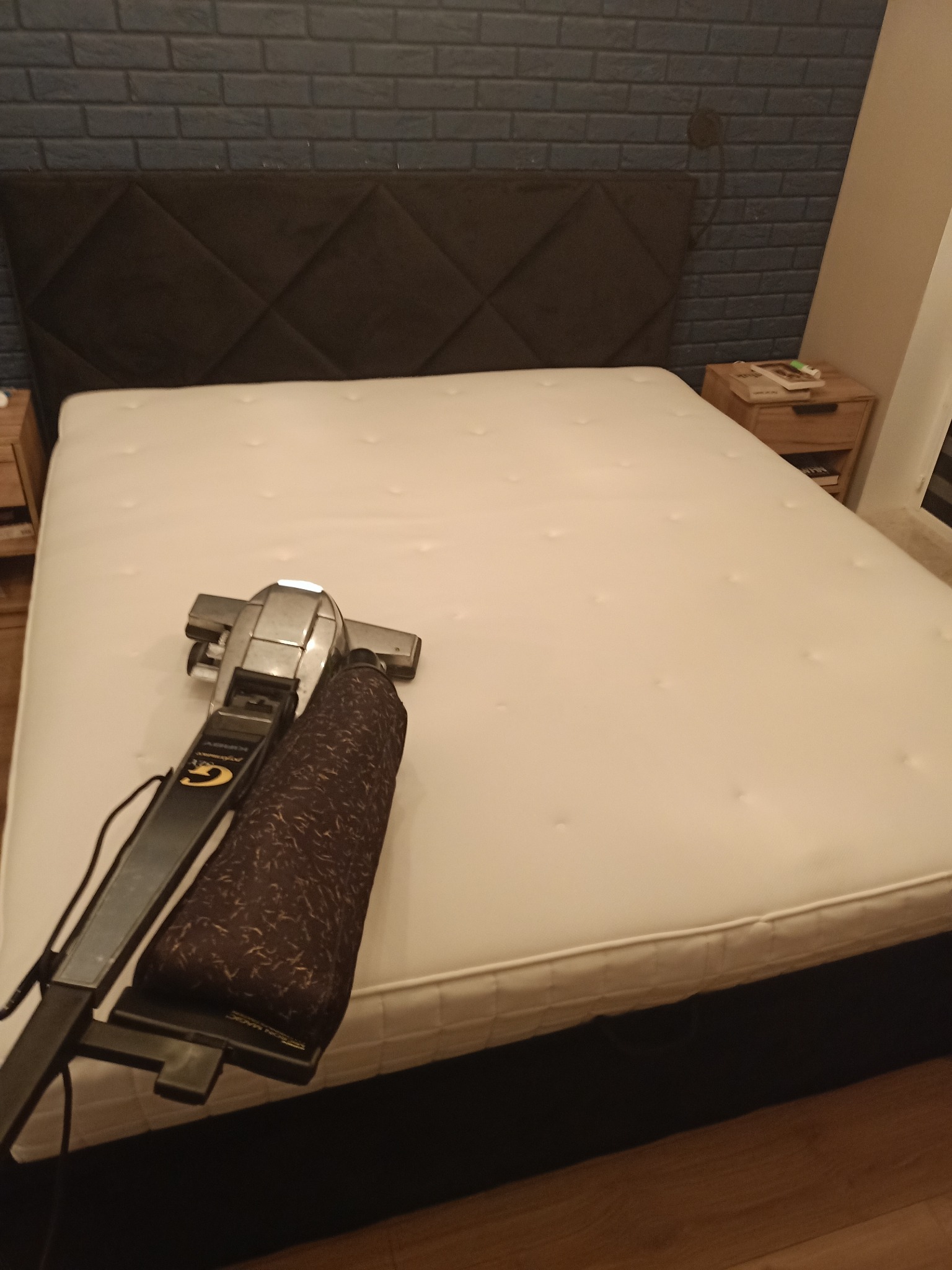 Czyszczenie materaca na łóżku za pomocą odkurzacza piorącego; widoczna tapicerowana rama łóżka i ceglana ściana w tle.