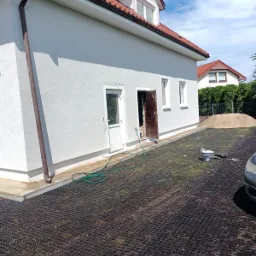 Świeżo pomalowana, jasna elewacja domu jednorodzinnego z czerwoną dachówką, widoczne narzędzia malarskie i wąż ogrodowy na ażurowym podłożu. Dom po remoncie.