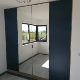 Nowoczesna szafa wnękowa z lustrzanymi drzwiami przesuwnymi i ciemnogranatowymi panelami bocznymi, widoczna w pustym pokoju z oknem i minimalistyczną lampą sufitową.