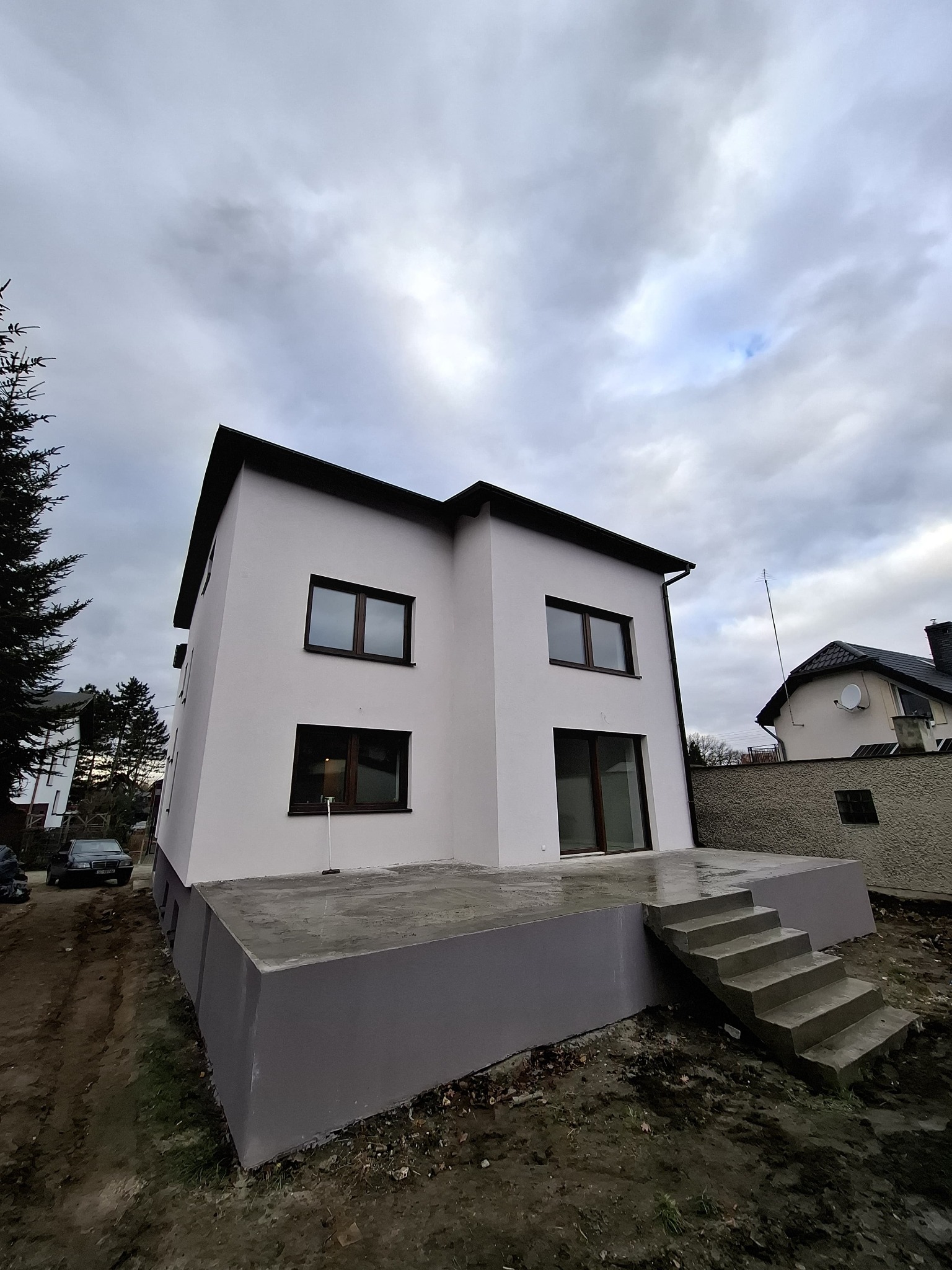 Nowoczesny, dwukondygnacyjny dom z białym tynkiem zewnętrznym i ciemnymi ramami okiennymi, widok z dołu na tle pochmurnego nieba, z widocznym betonowym tarasem i schodami wejściowymi.