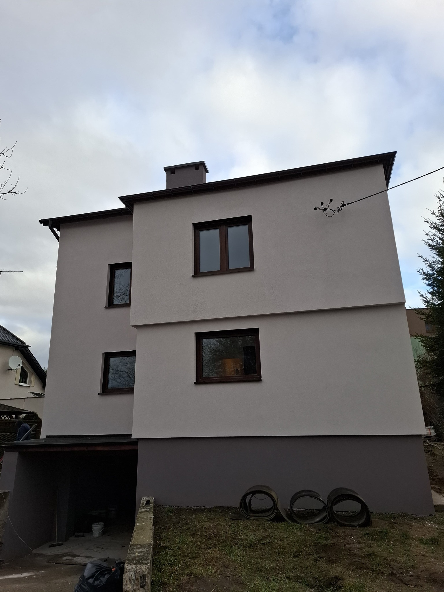 Trzykondygnacyjny dom z jasnoszarą elewacją, ciemnobrązowymi oknami i ciemnoszarą dolną częścią, z widocznymi trzema zwiniętymi rolkami papy na trawniku przed budynkiem.