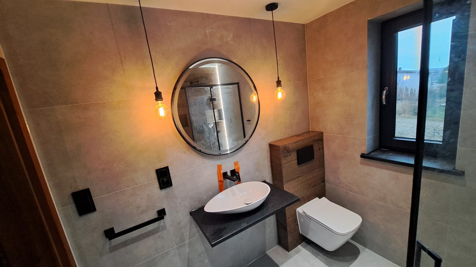 Nowoczesna łazienka z beżowymi płytkami, owalną umywalką nablatową na czarnym blacie, okrągłym lustrem i wiszącą toaletą z obudową z drewna.