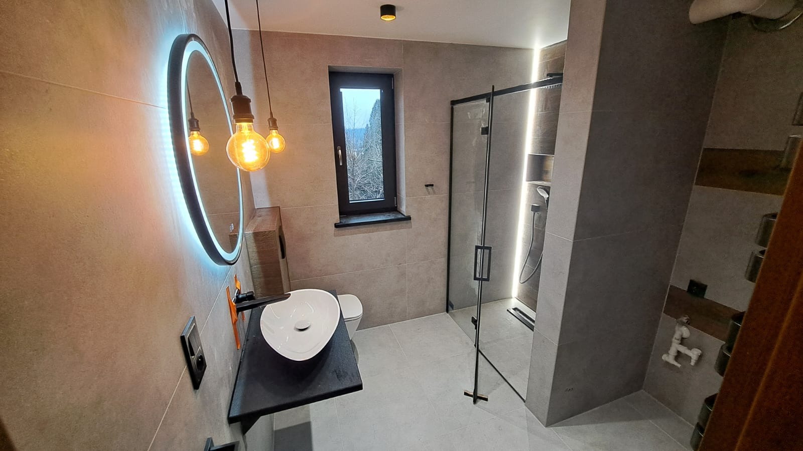 Nowoczesna łazienka z szarymi płytkami, okrągłym lustrem z podświetleniem LED, wiszącą umywalką na czarnym blacie i kabiną prysznicową typu walk-in z czarnymi profilami, oświetlona trzema żarówkami...