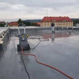 Czyszczenie dachu płaskiego za pomocą myjki ciśnieniowej; widoczny pracownik w kombinezonie, duży budynek w tle.